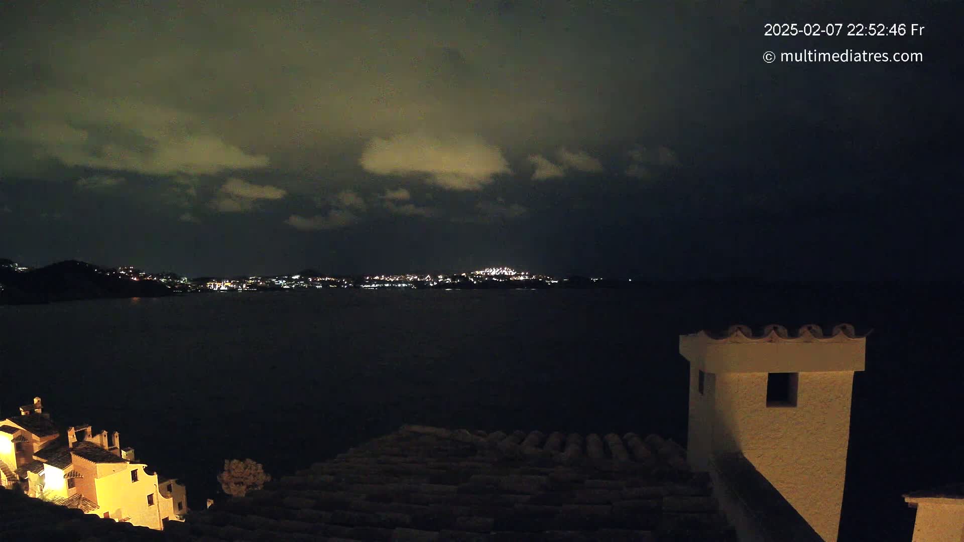 A nighttime view across a dark body of water shows a distant, faintly lit city under a cloudy sky, with a portion of a building and its roof in the foreground.
