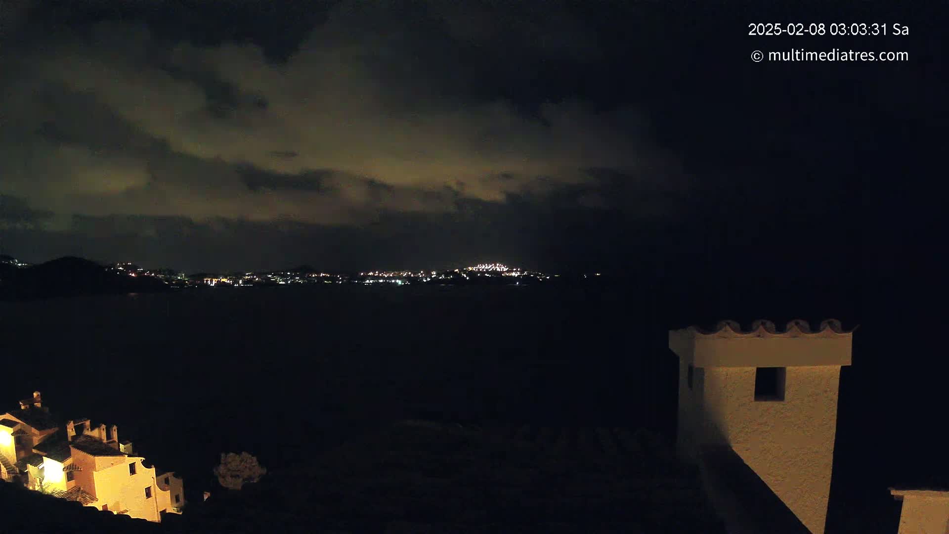 A nighttime view of a city across a body of water under a cloudy sky, partially obscured by a building and chimney in the foreground.