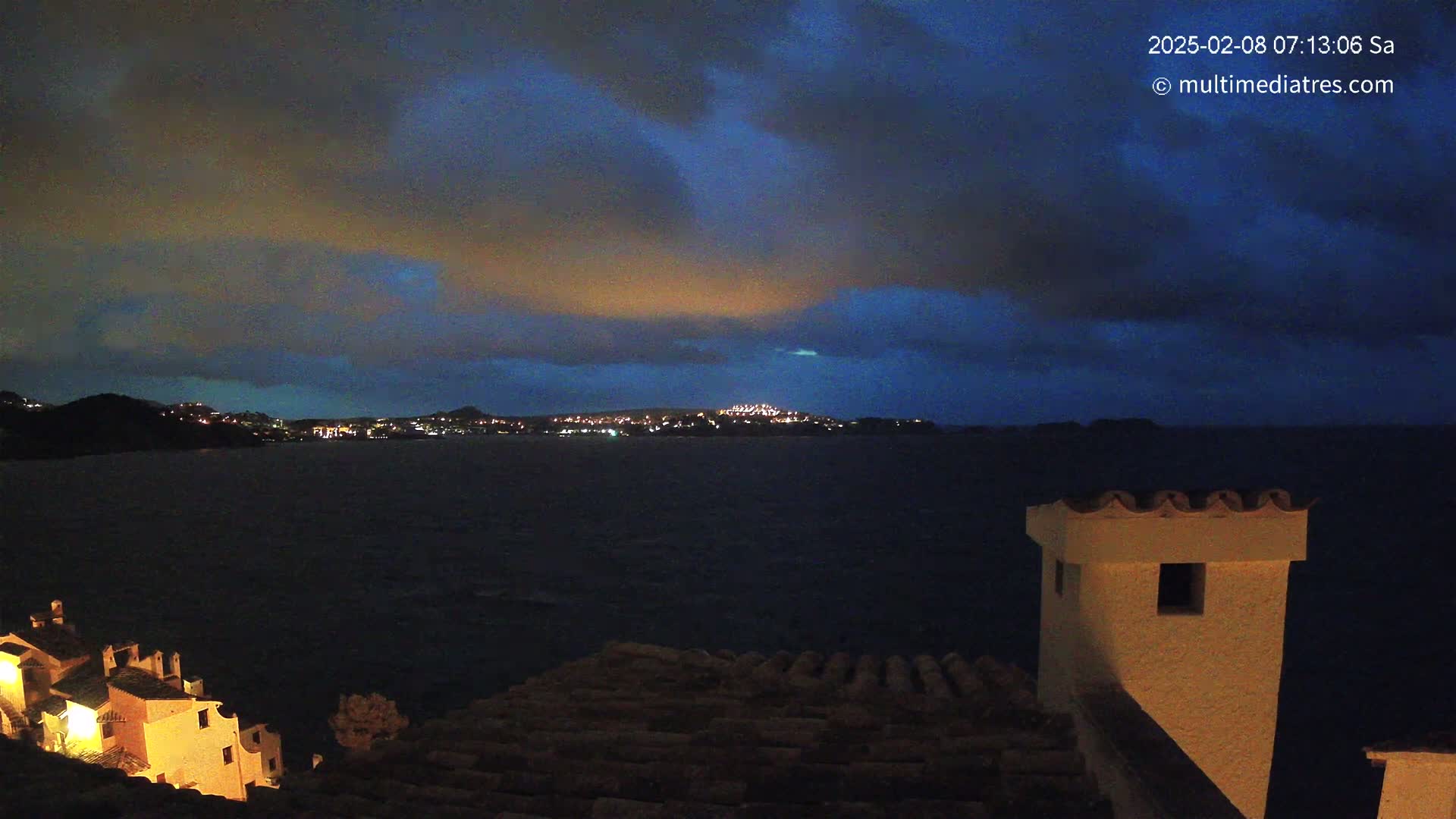 A nighttime coastal scene shows a dark body of water with a distant, lit town under a mostly cloudy, dark blue sky.