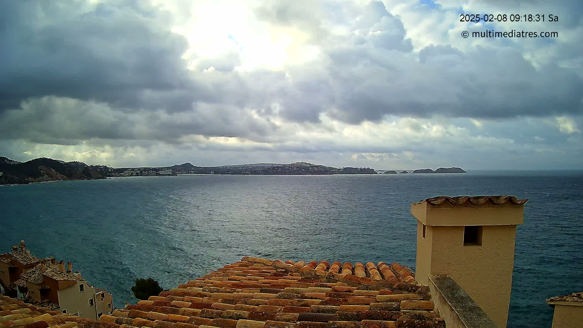 A view from a rooftop shows a calm sea under a cloudy sky, with distant hills and a coastline visible.