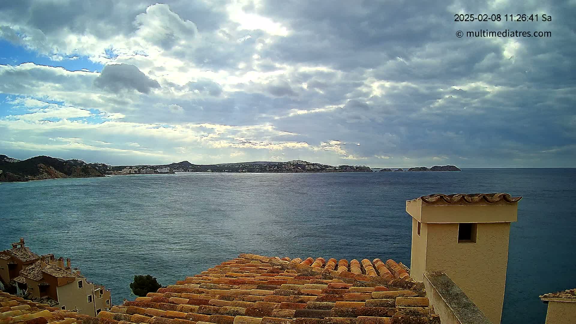 A partly cloudy sky overlooks a calm sea with a distant coastline and buildings, viewed from a rooftop with terracotta tiles and a small, light-beige chimney.