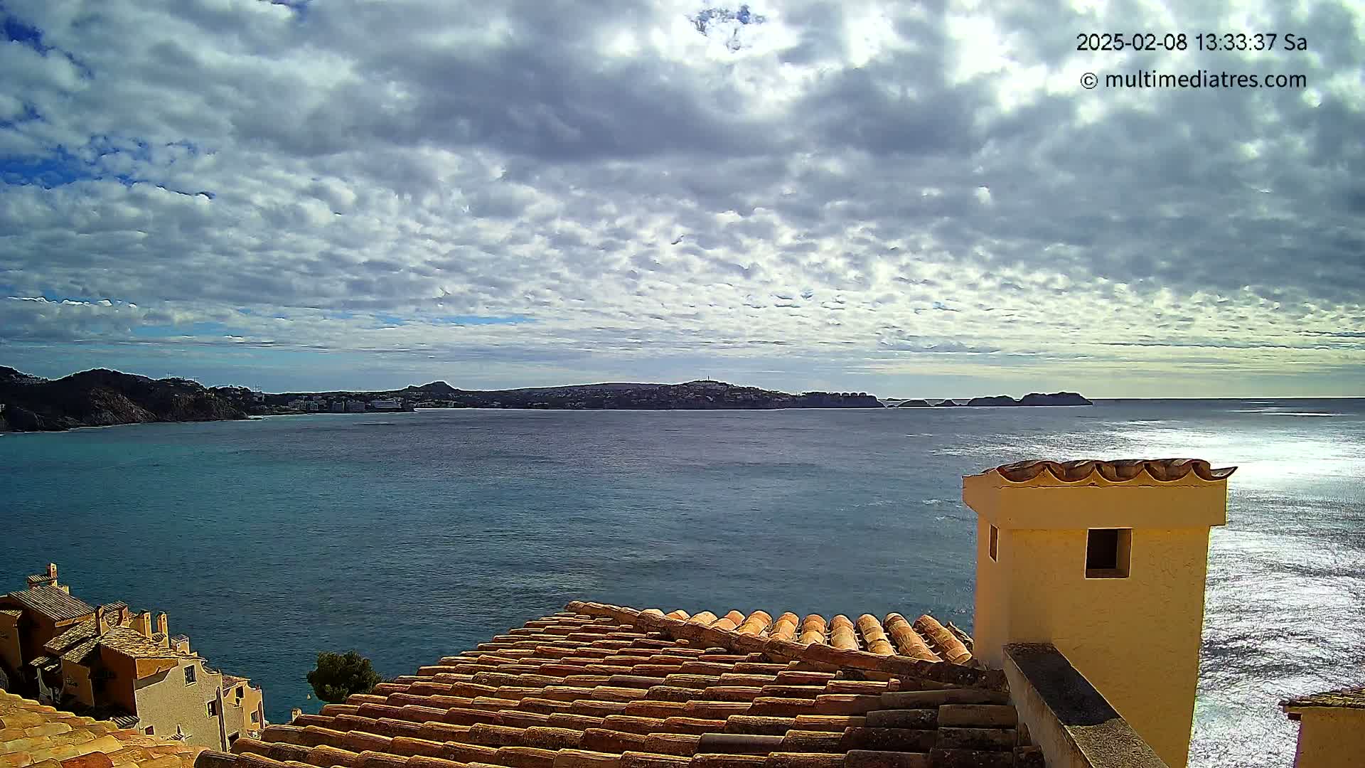 A partly cloudy sky overlooks a calm sea with distant hills and a building's terracotta roof and chimney in the foreground.