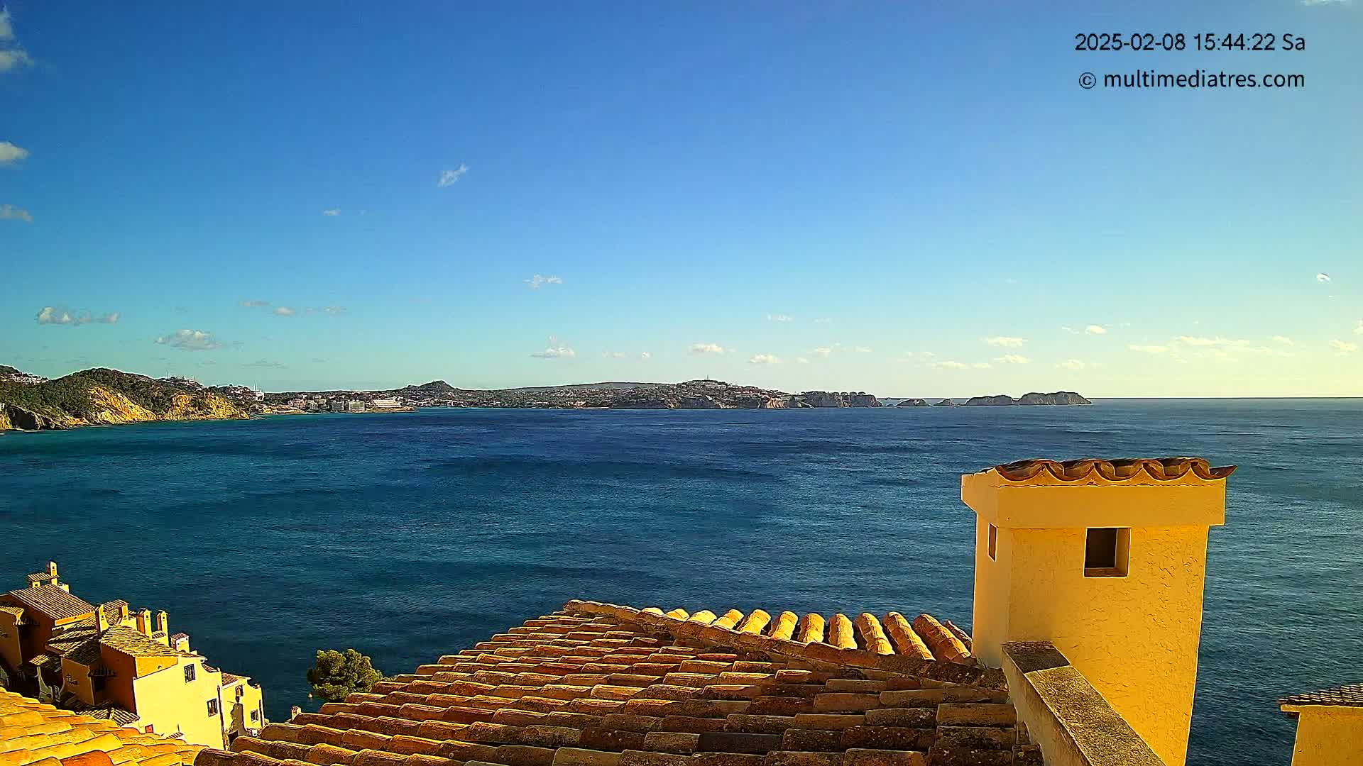 A clear blue sky overlooks a calm blue sea with a distant coastline, viewed from a rooftop with orange-tiled roofs and a yellow chimney.