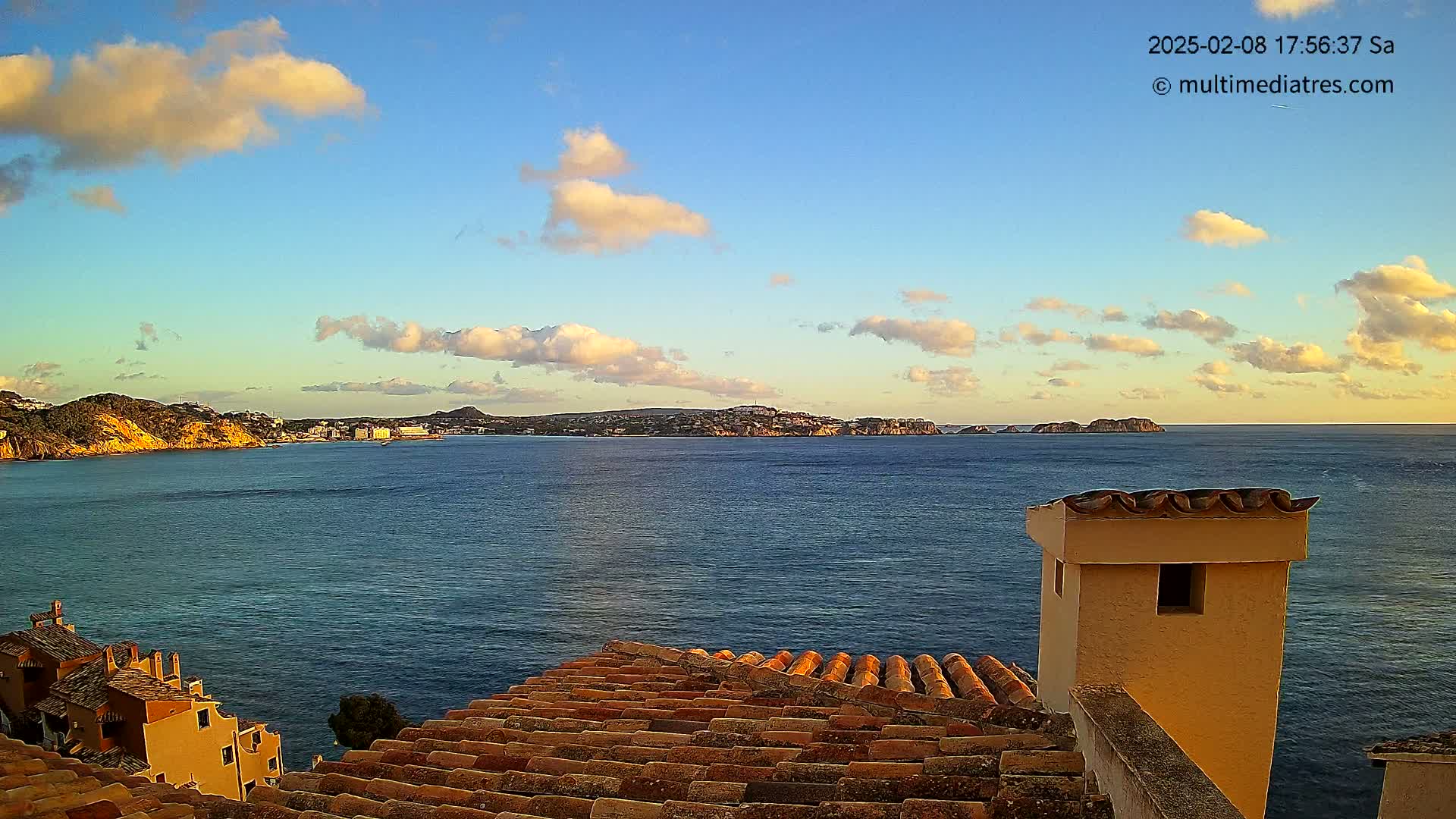 A partly cloudy sunset over a calm ocean viewed from a tiled rooftop, with a coastal town and islands in the distance.
