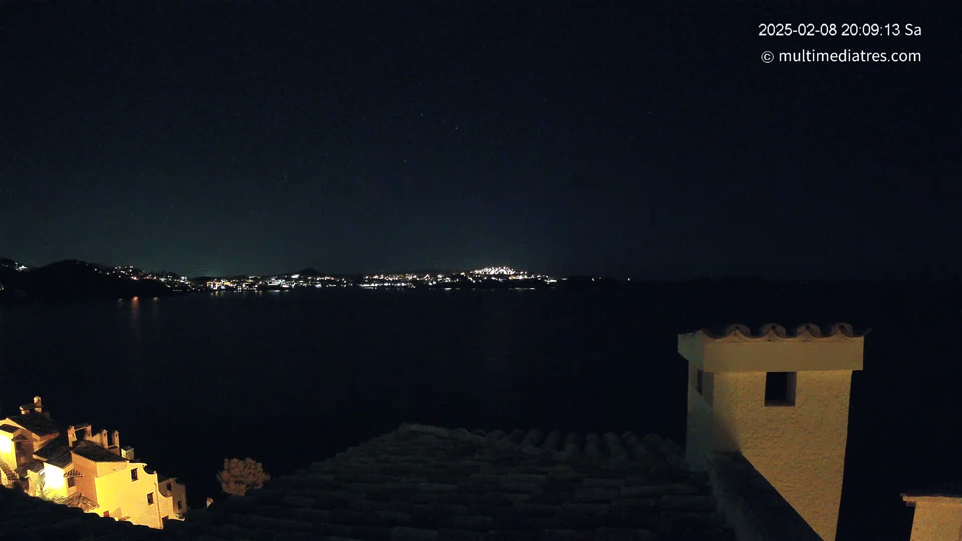 A nighttime view of a city's lights across a dark body of water, seen from a vantage point on a rooftop with a nearby building illuminated.