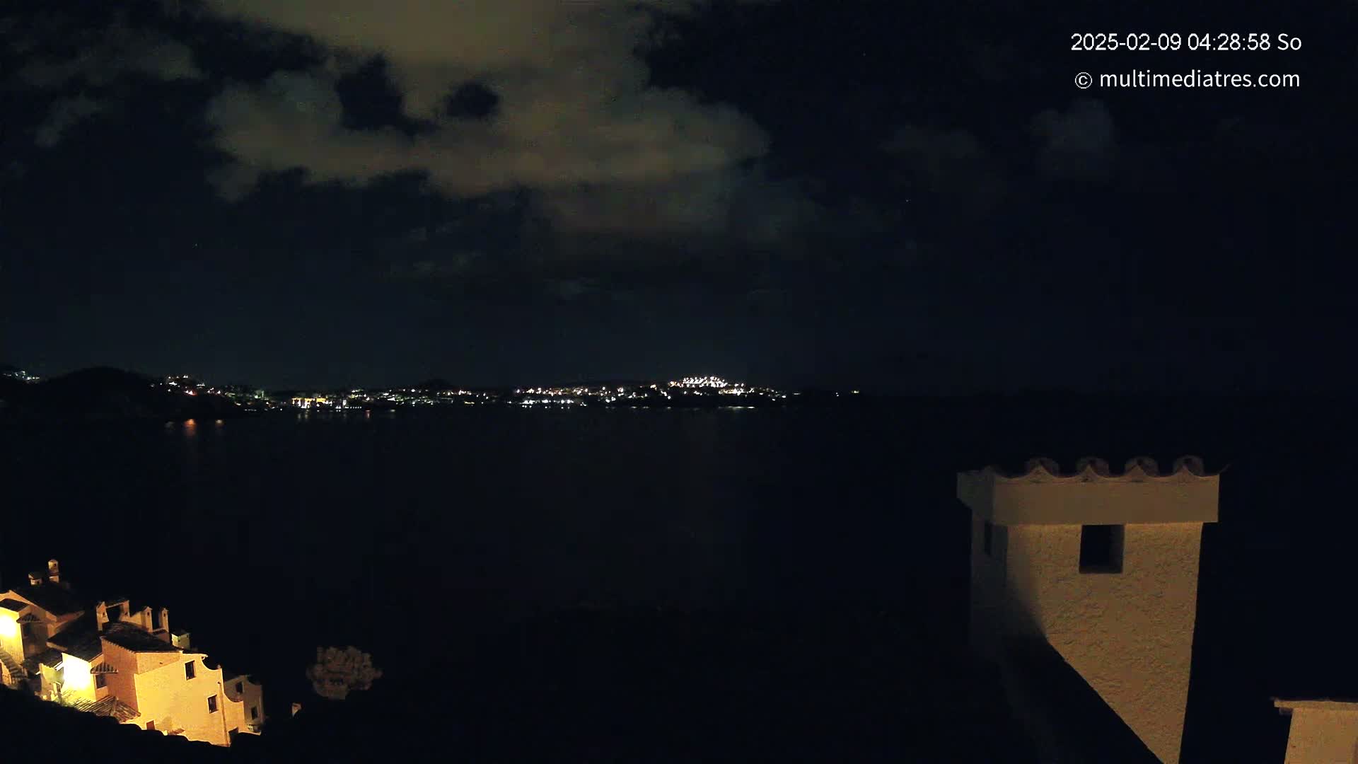 A nighttime view of a city's lights across a dark body of water, under a partly cloudy sky, with a building's illuminated chimney in the foreground.
