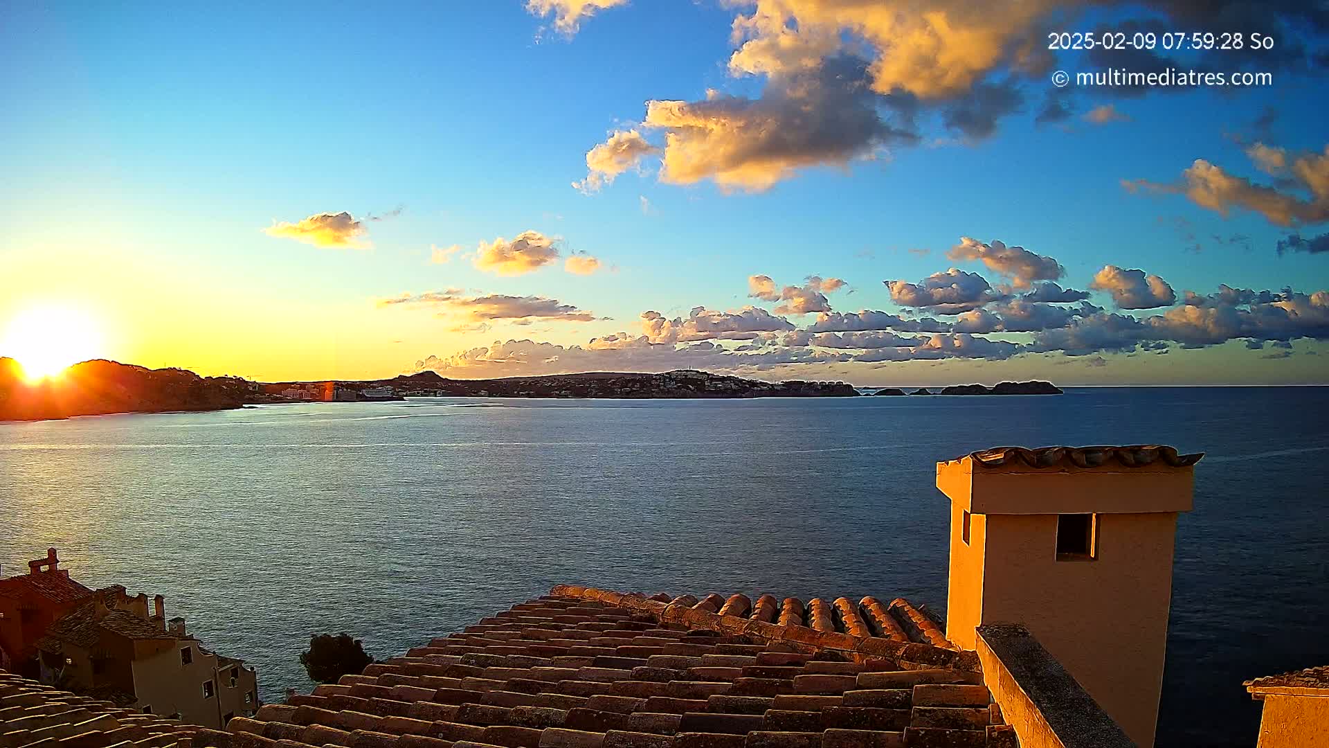 A sunrise over a calm ocean with a distant coastline and a section of a tiled roof and chimney in the foreground, under a partly cloudy sky.