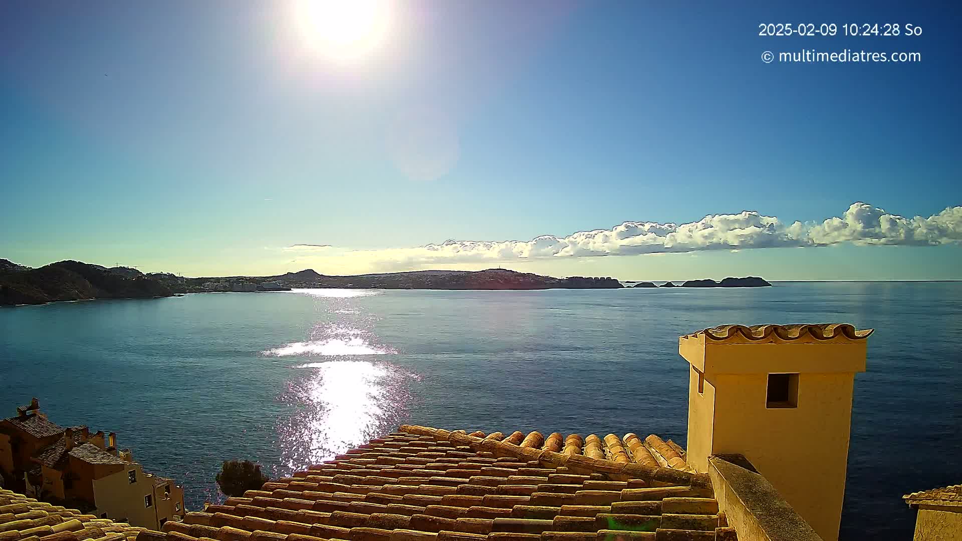 A sunny day reveals a coastal scene with a calm sea reflecting sunlight, viewed from a terracotta roof overlooking a town nestled against hills and small islands in the distance.