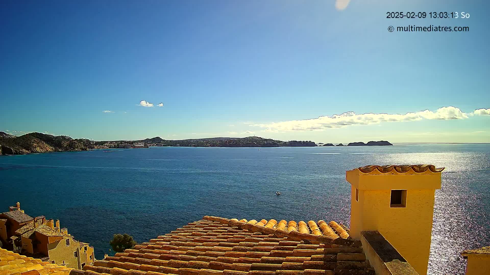 A sunny day overlooks a calm blue sea, with distant hills and islands visible from a terracotta roof and yellow chimney.