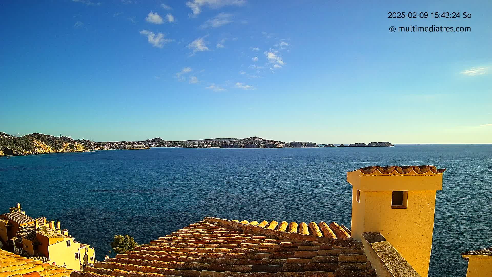 A view from a terracotta roof shows a calm, blue sea extending to a distant shoreline under a mostly clear blue sky with a few clouds.