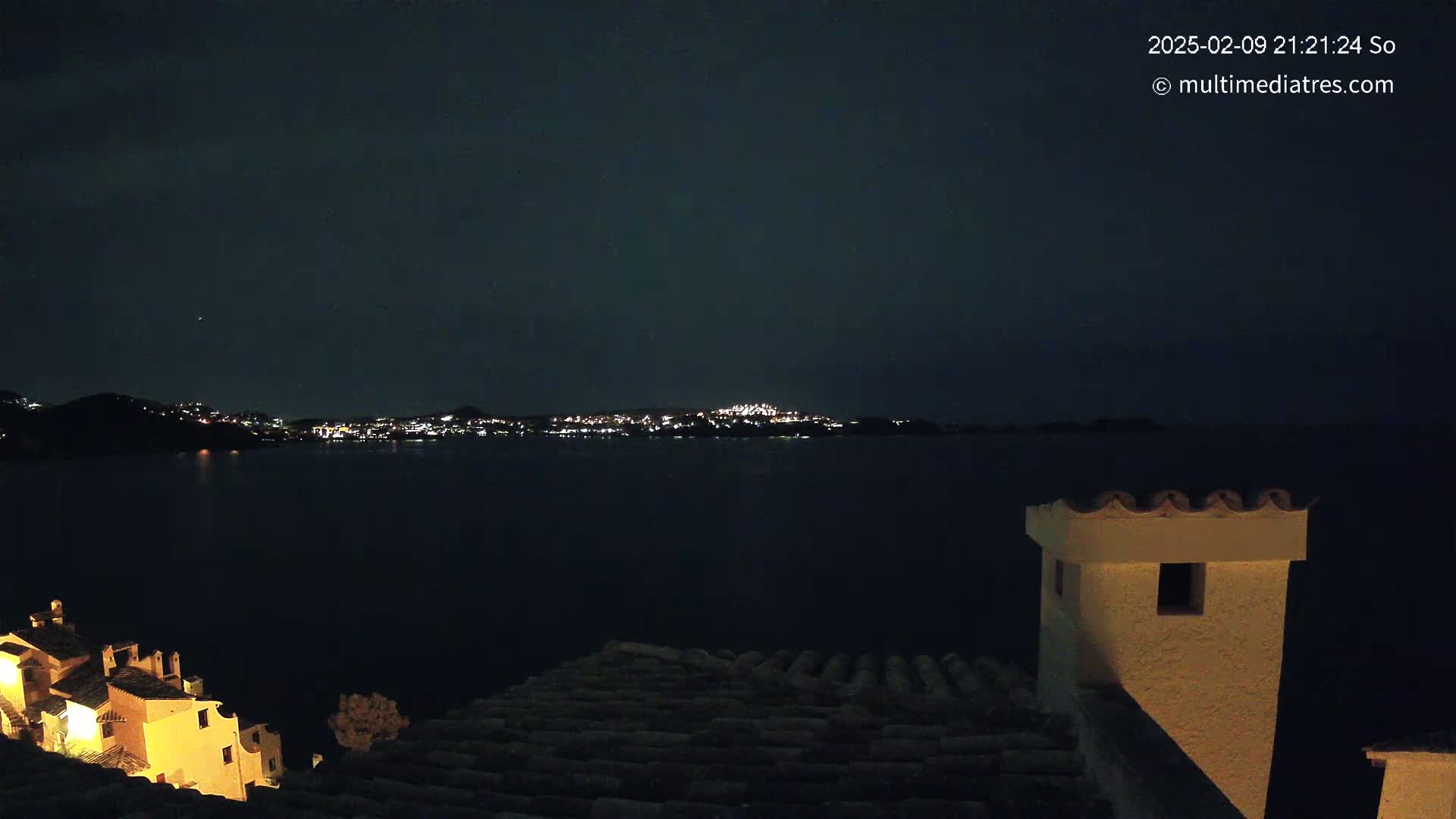 A nighttime view from a rooftop shows a dark body of water reflecting the lights of a distant town under a clear, dark sky.