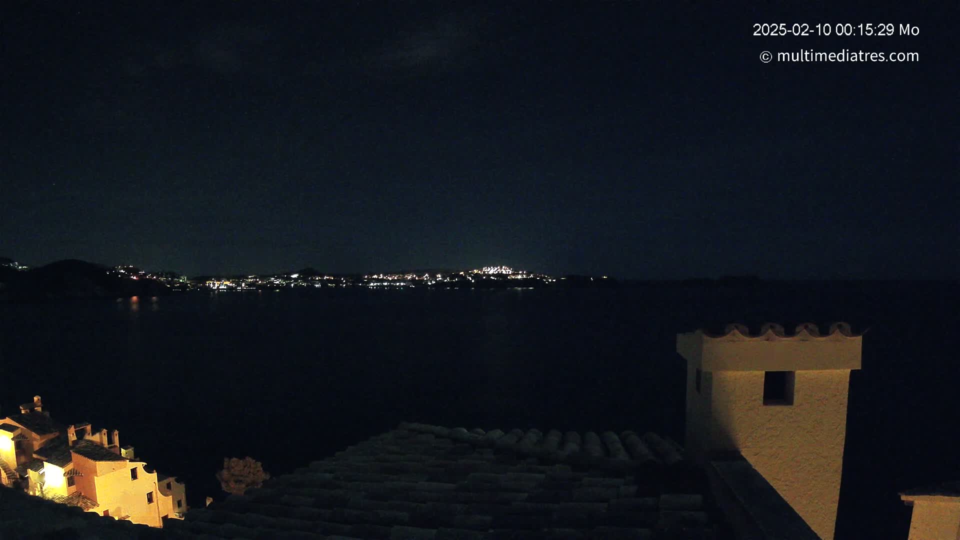 A nighttime view from a rooftop shows a dark body of water with a distant city skyline illuminated, under a mostly clear night sky.