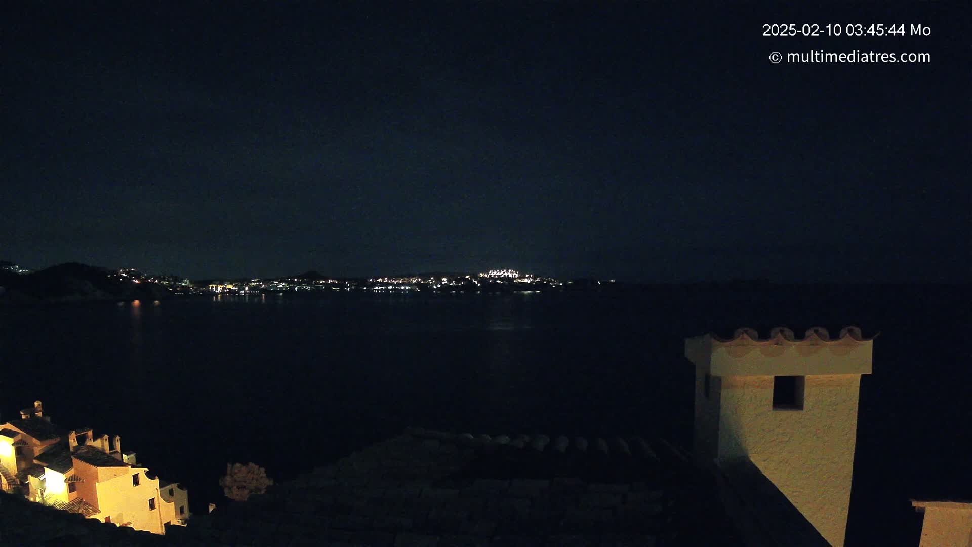 A nighttime view of a dark body of water with a city illuminated on the far shore, seen from a vantage point overlooking buildings with lit windows.