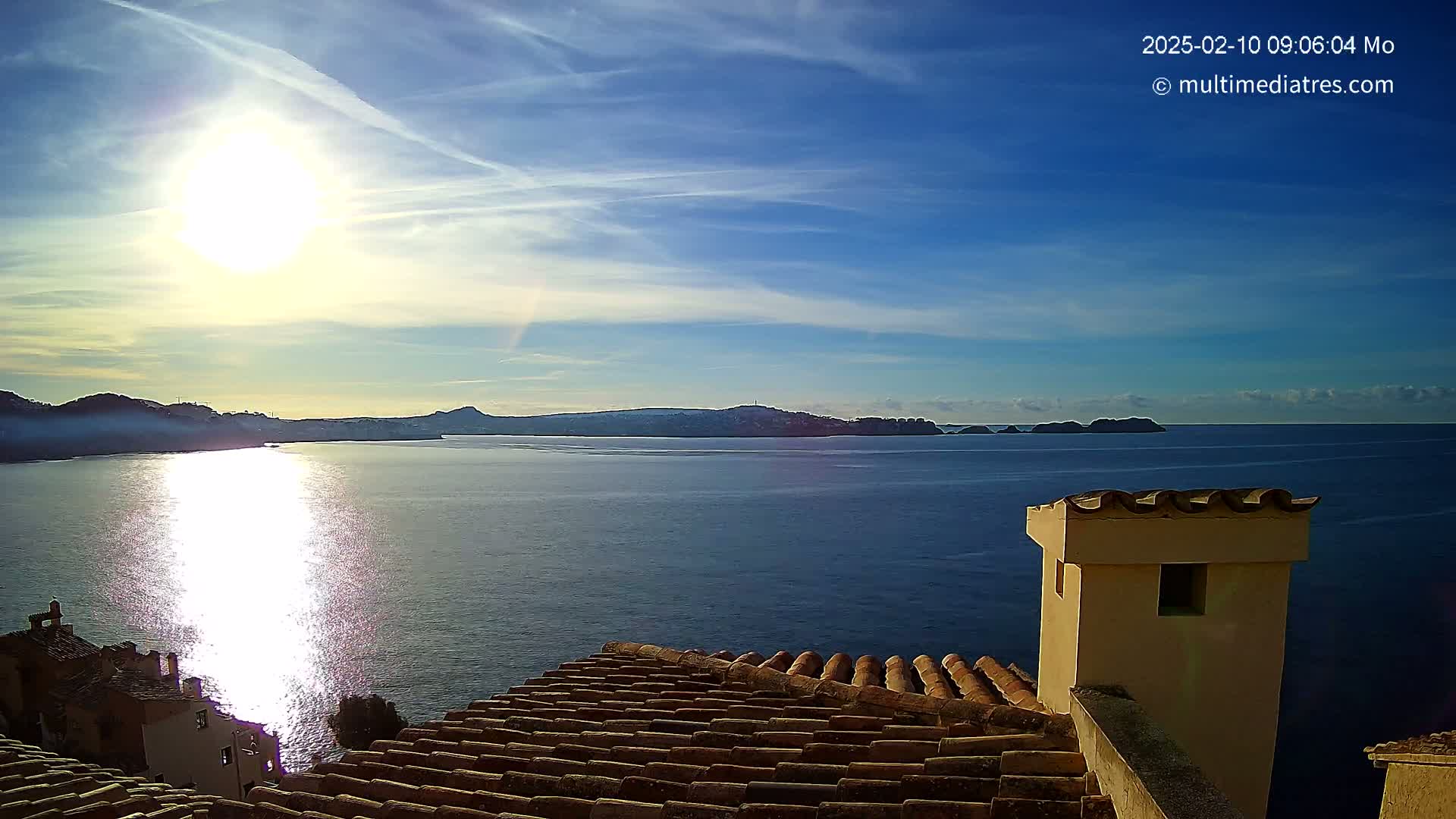 A bright sun shines over a calm sea, reflecting on the water, with distant hills and islands visible under a mostly clear blue sky, viewed from a tiled roof with a tan chimney.
