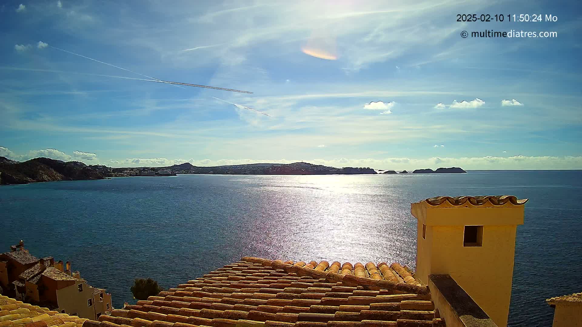 A sunny day with a few clouds shows a coastal view of a calm blue sea with islands in the distance, seen from a rooftop with orange-brown tiles.