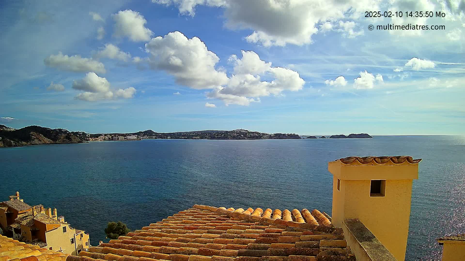 A partly cloudy blue sky overlooks a calm, deep blue sea with a distant coastline and buildings, viewed from a terracotta roof with a pale yellow chimney.