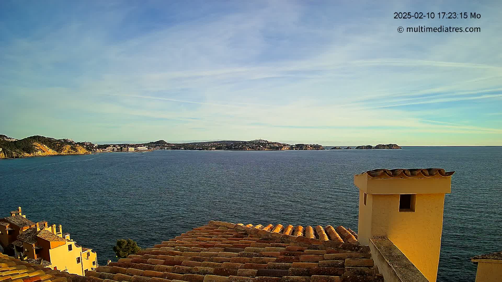 A coastal town is seen from a rooftop, overlooking a calm sea under a mostly clear sky.