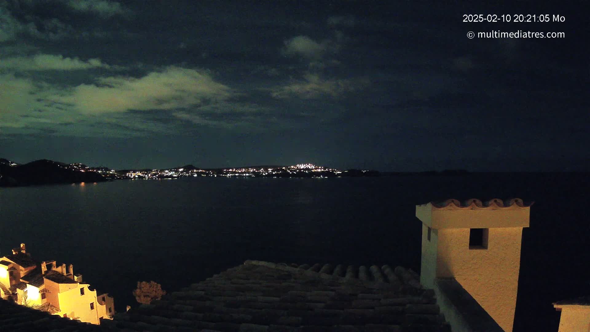 A nighttime view of a dark body of water with illuminated buildings across the water and a portion of a building with a chimney in the foreground under a cloudy sky.