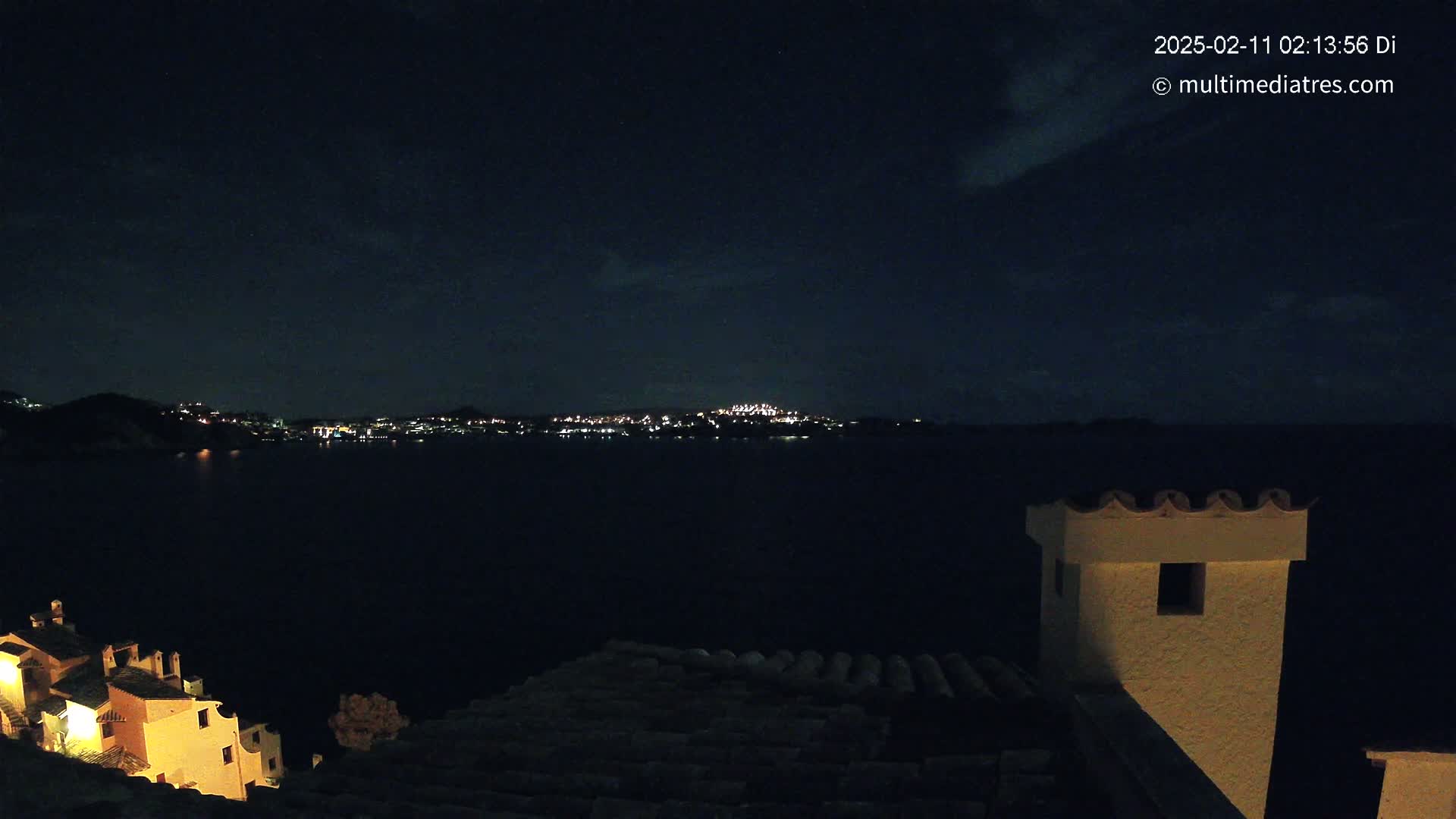 A nighttime view from a rooftop shows a dark body of water with a distant, illuminated town across it, under a mostly clear night sky.