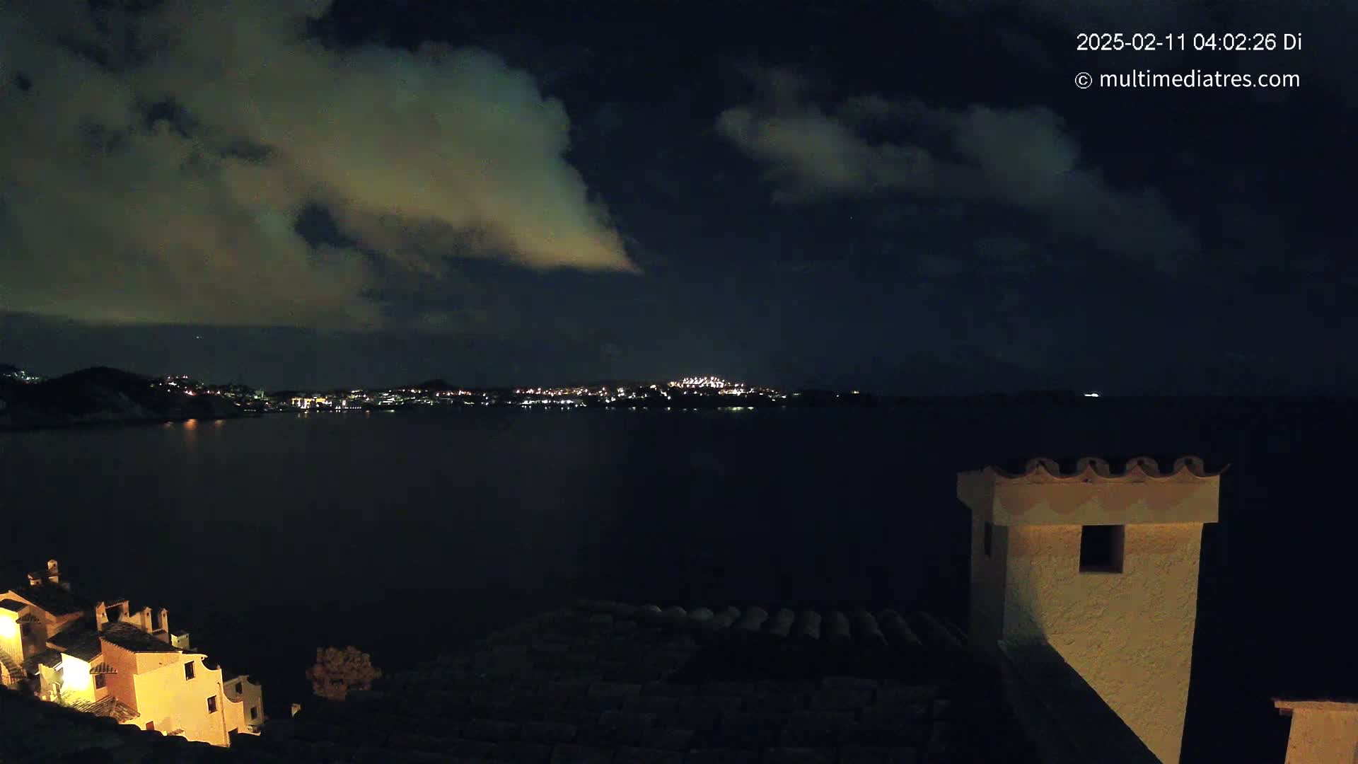 A nighttime view of a city across a dark body of water, under a partly cloudy sky, seen from a vantage point on a hillside with buildings illuminated.