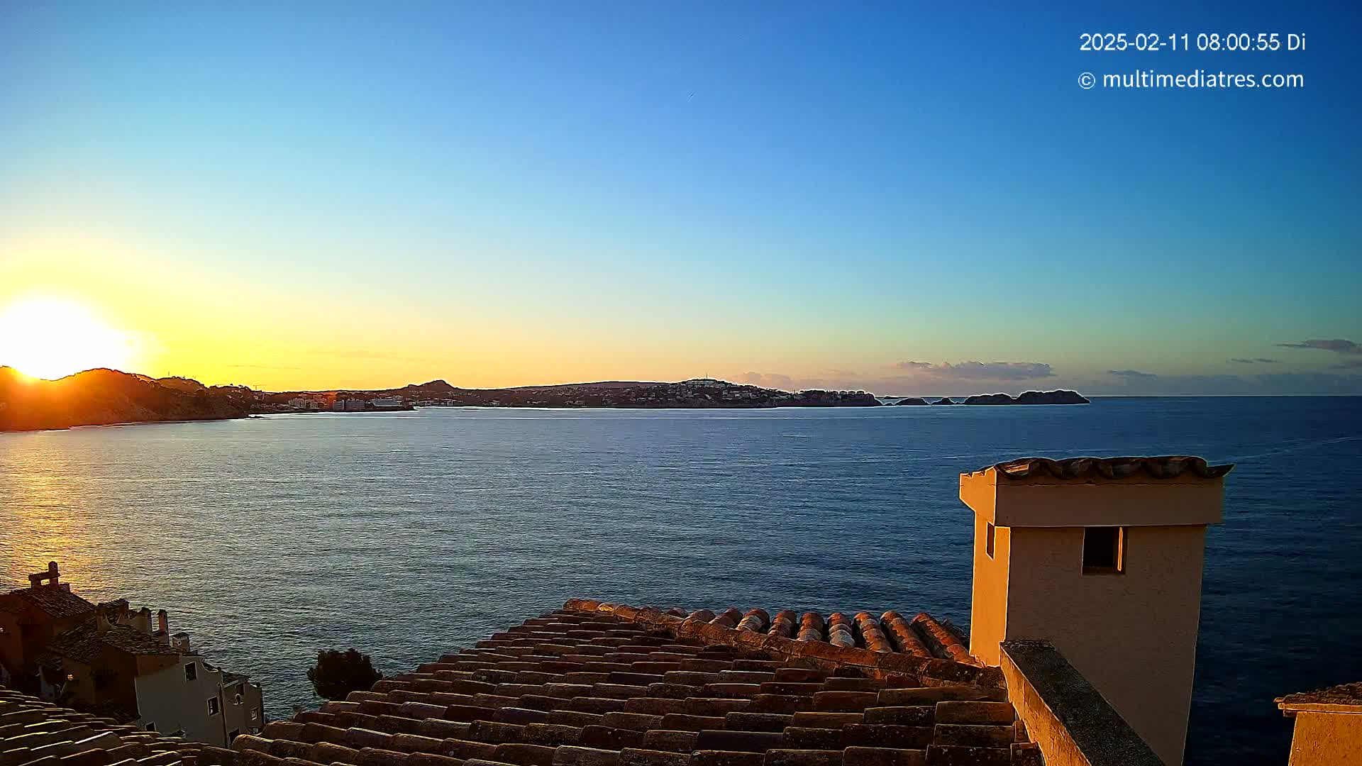 A coastal sunrise scene viewed from a tiled rooftop, showcasing calm waters and a distant landmass bathed in the golden light of the rising sun.
