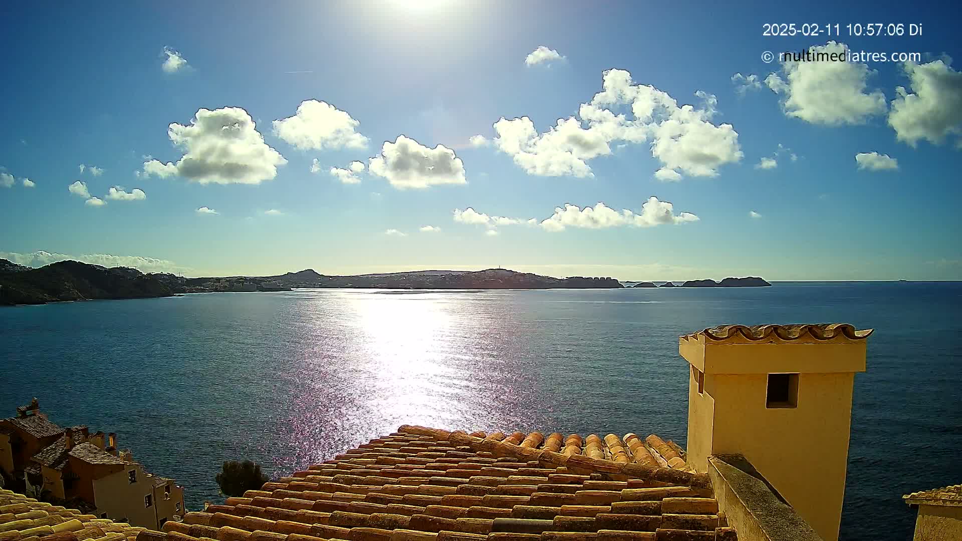 A sunny day with scattered clouds overlooks a calm sea with distant hills and islands, as seen from a tiled rooftop with a small building nearby.