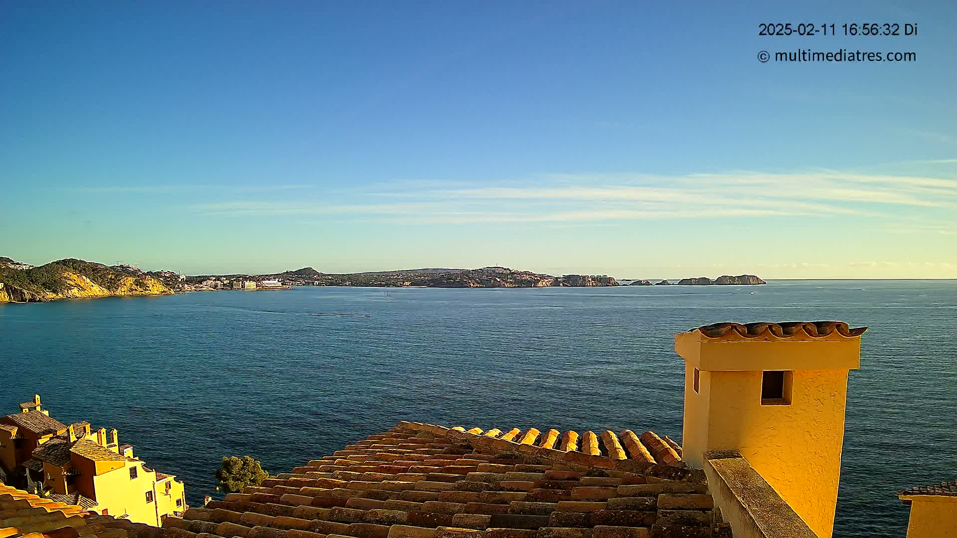 A coastal view on a clear, sunny day shows a calm sea extending to a distant shoreline, with a portion of a terracotta roof and a yellow chimney in the foreground.