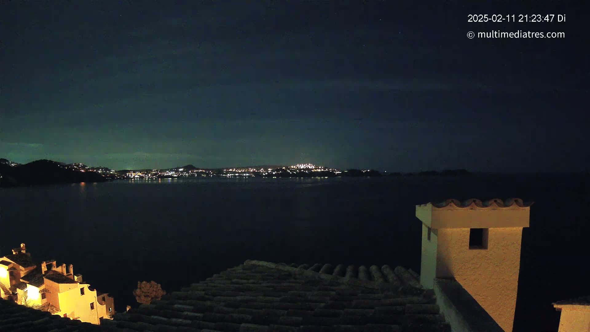 A nighttime view from a rooftop shows a dark body of water reflecting the lights of a city across the water, under a clear, dark sky.