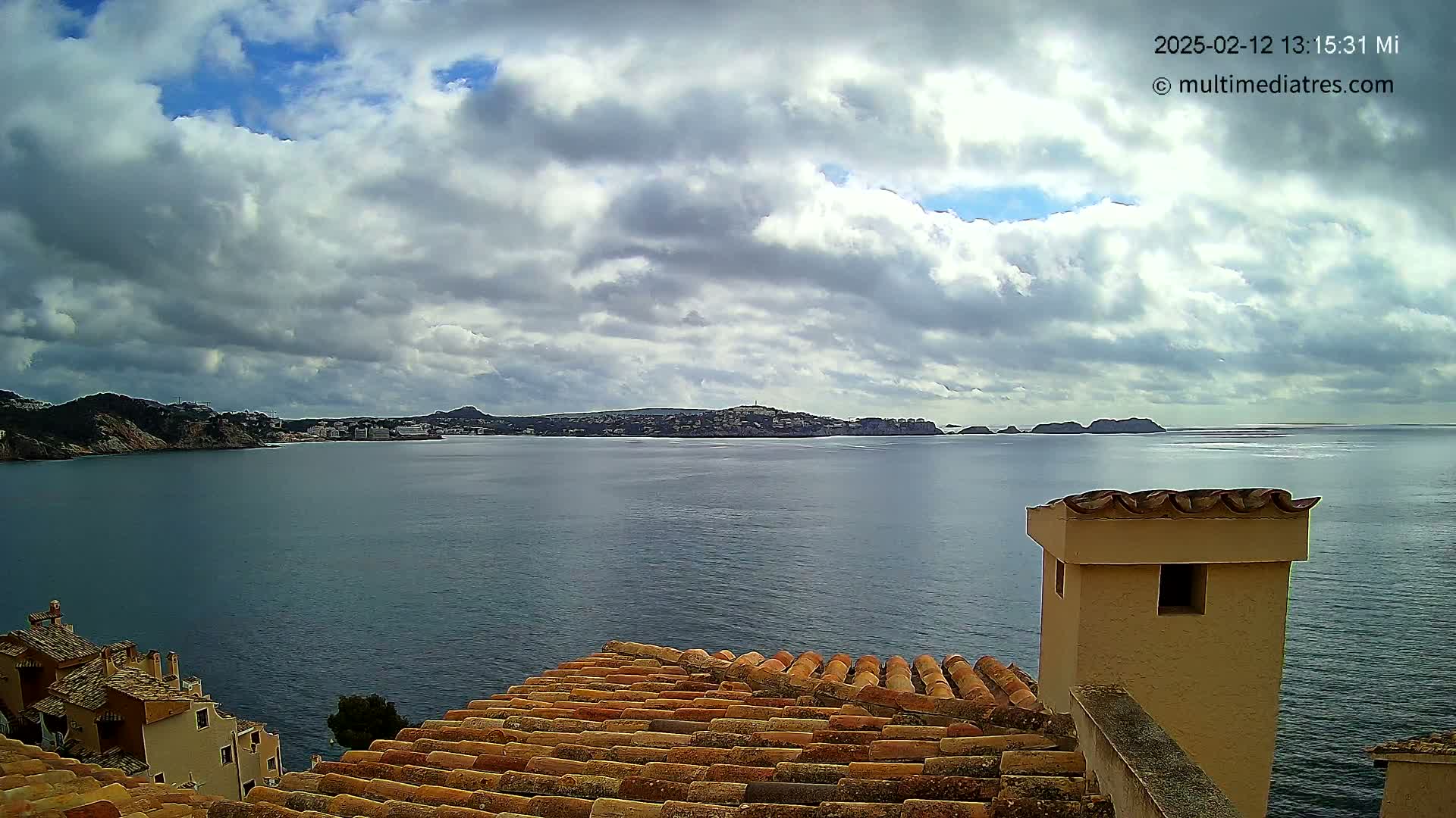 A coastal view on a partly cloudy day shows a calm body of water with a distant shoreline and buildings, seen from a rooftop with terracotta tiles.
