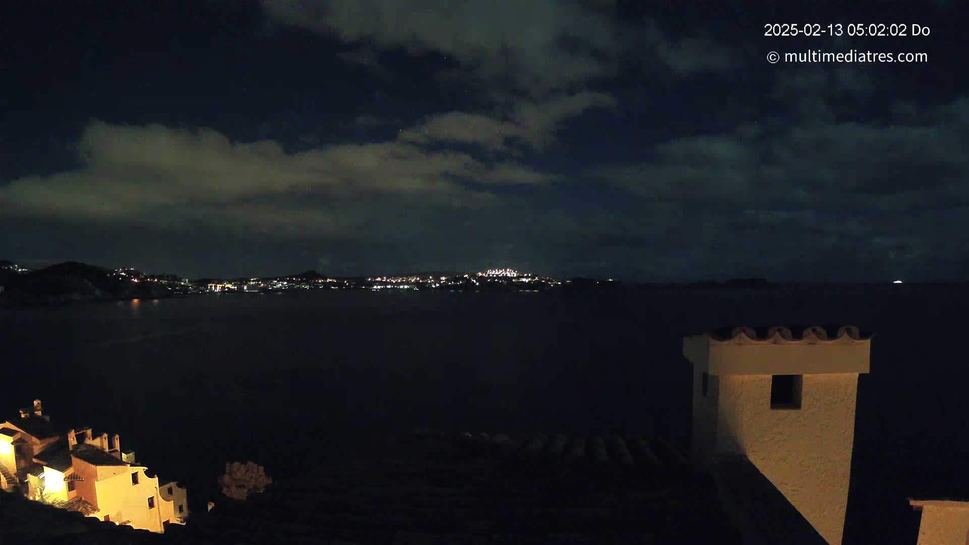 A nighttime coastal scene shows a dark body of water separating a village with illuminated buildings from a distant city skyline under a cloudy night sky.
