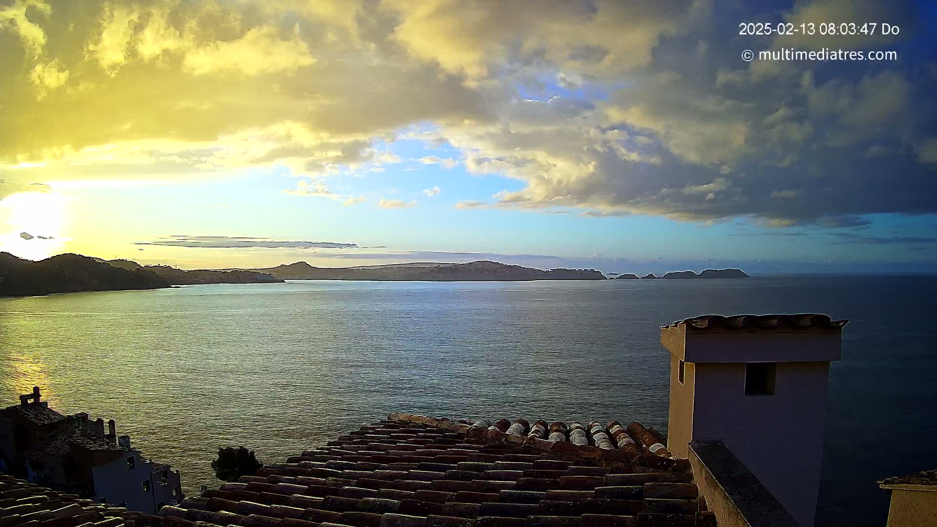 A sunrise over a calm ocean with distant islands and hills, viewed from a rooftop with terracotta tiles.
