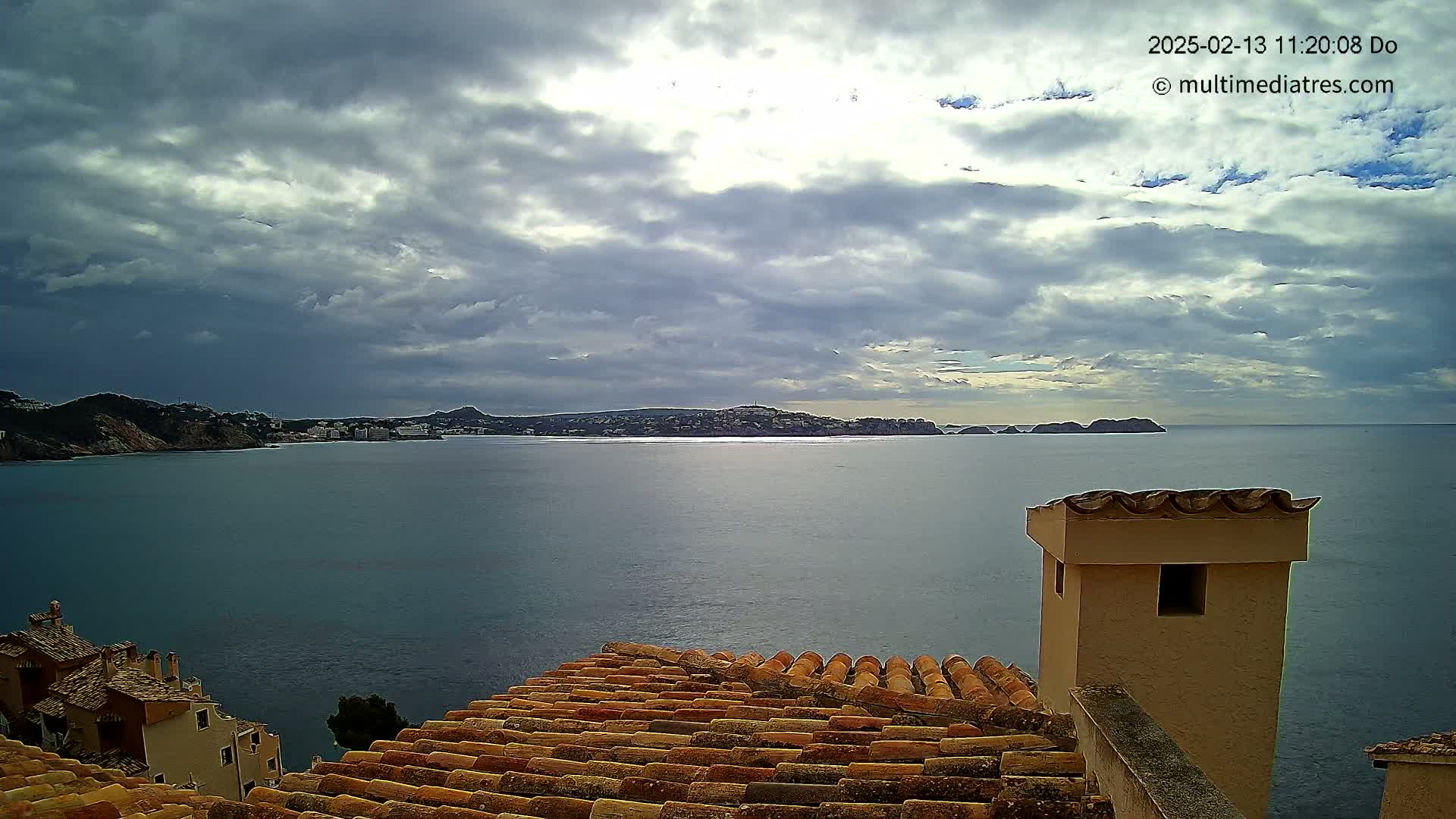 A partly cloudy sky overlooks a calm sea with a distant town nestled between hills, viewed from a rooftop with terracotta tiles and a tan chimney.