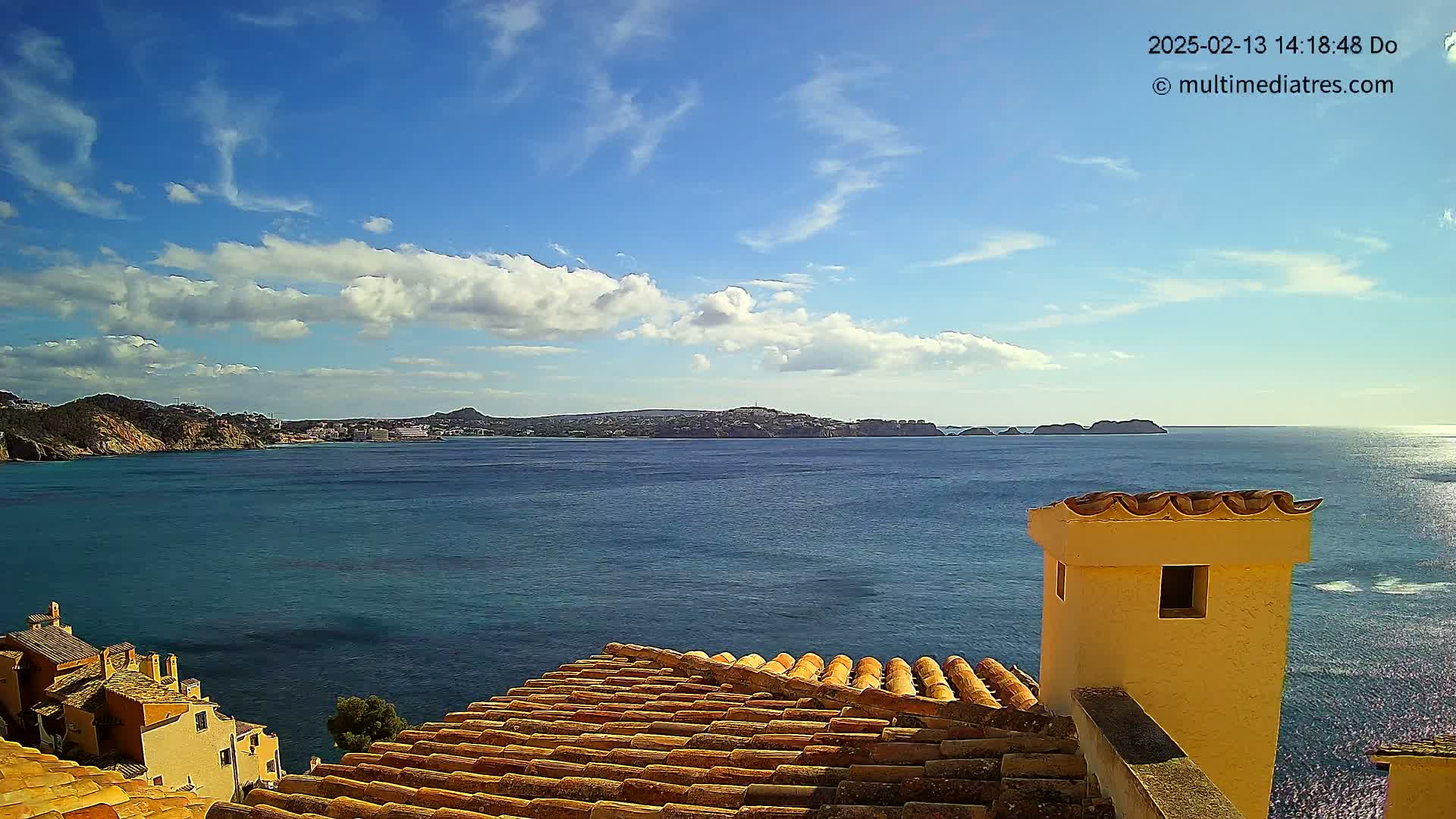 A partly cloudy blue sky overlooks a calm sea with a distant coastline, viewed from a rooftop with terracotta tiles and a yellow chimney.