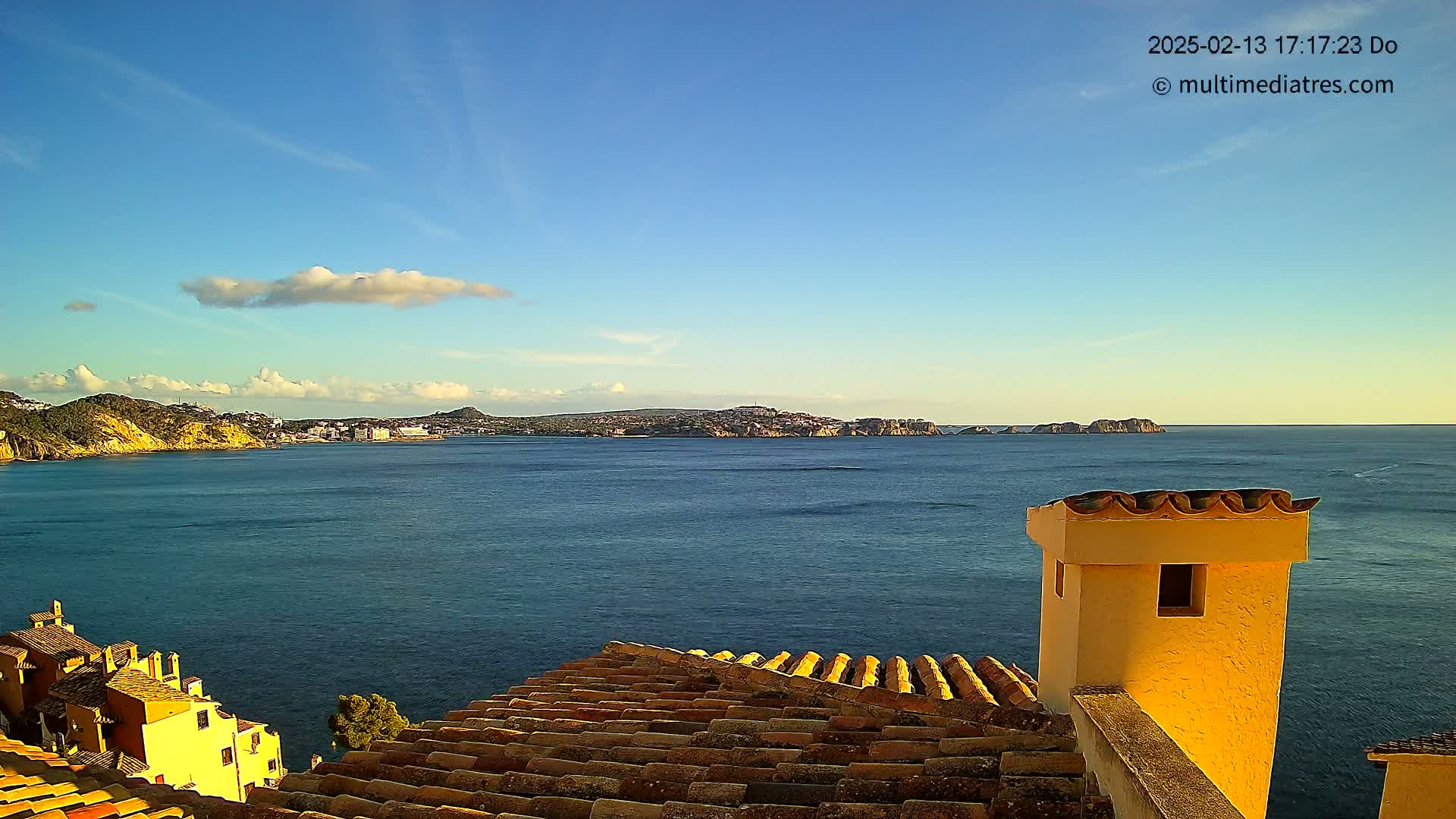 A clear blue sky with a few clouds overlooks a calm blue sea, with a distant town nestled between rocky headlands, viewed from a tiled rooftop with a yellow chimney.