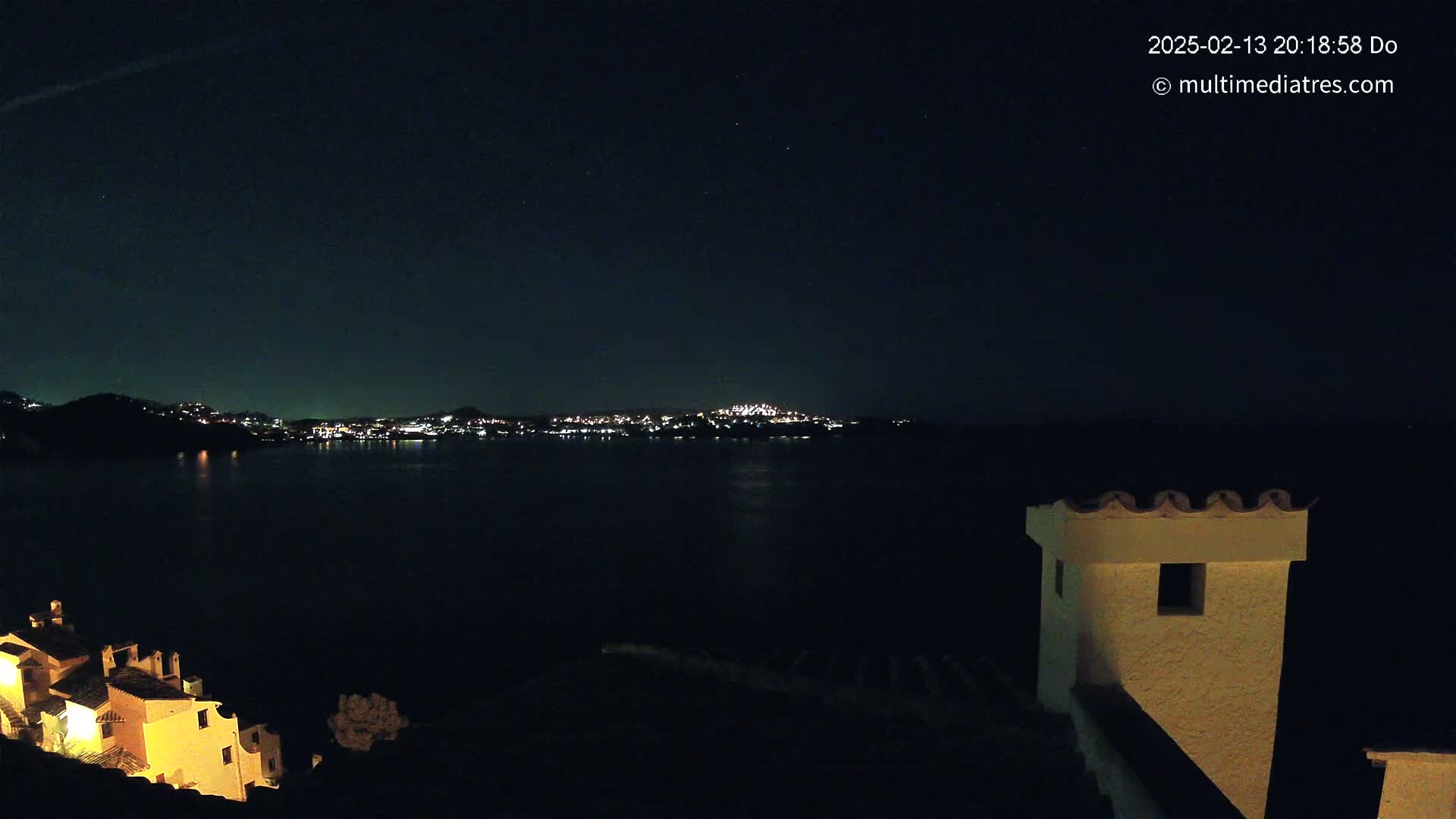 A nighttime view of a calm body of water with a city's lights on the opposite shore, partially obscured by hills, with a building's illuminated structure in the foreground under a clear, starry sky.