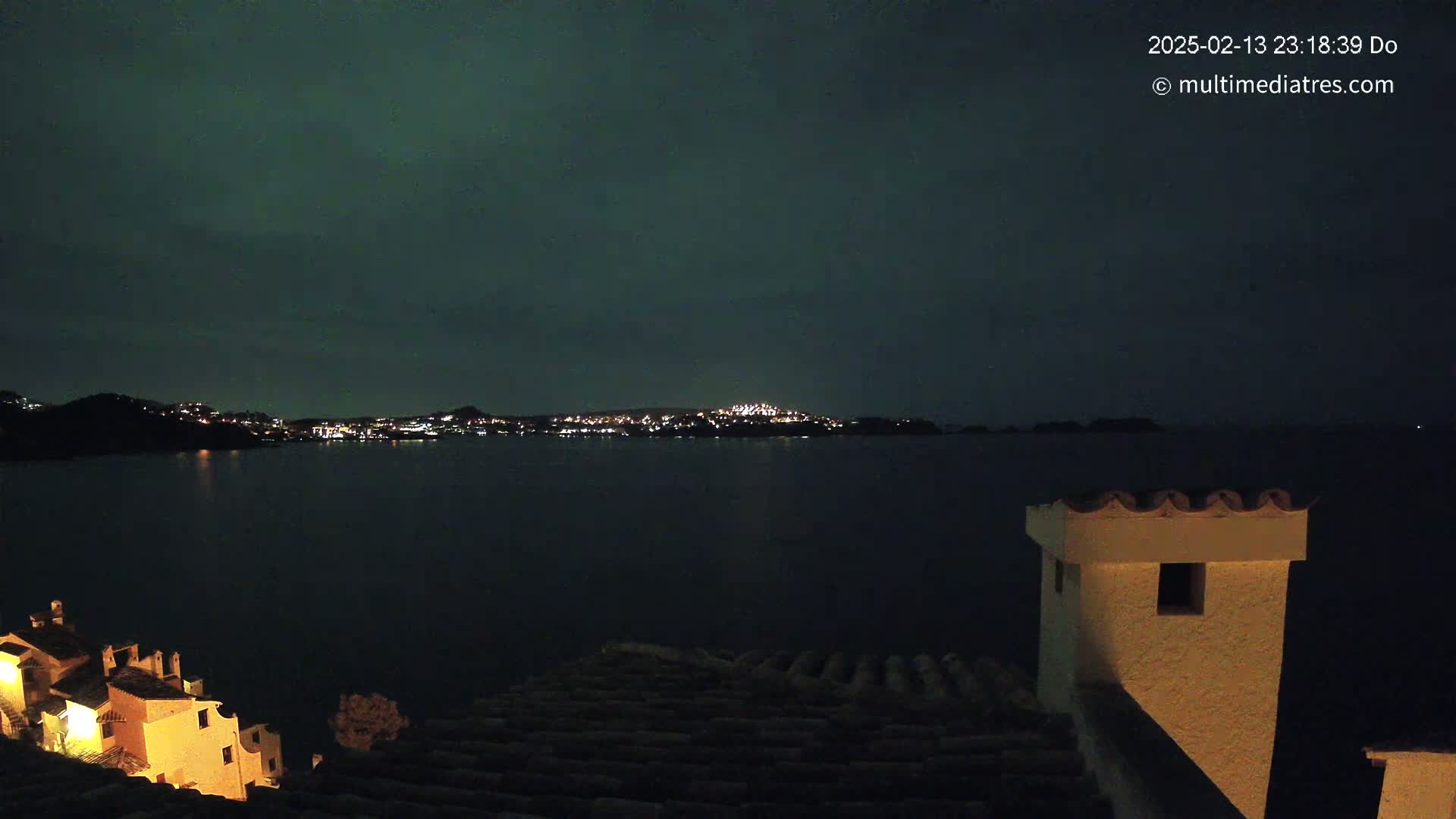 A nighttime view of a calm body of water with a distant, illuminated town across the water, under a dark, slightly greenish sky.
