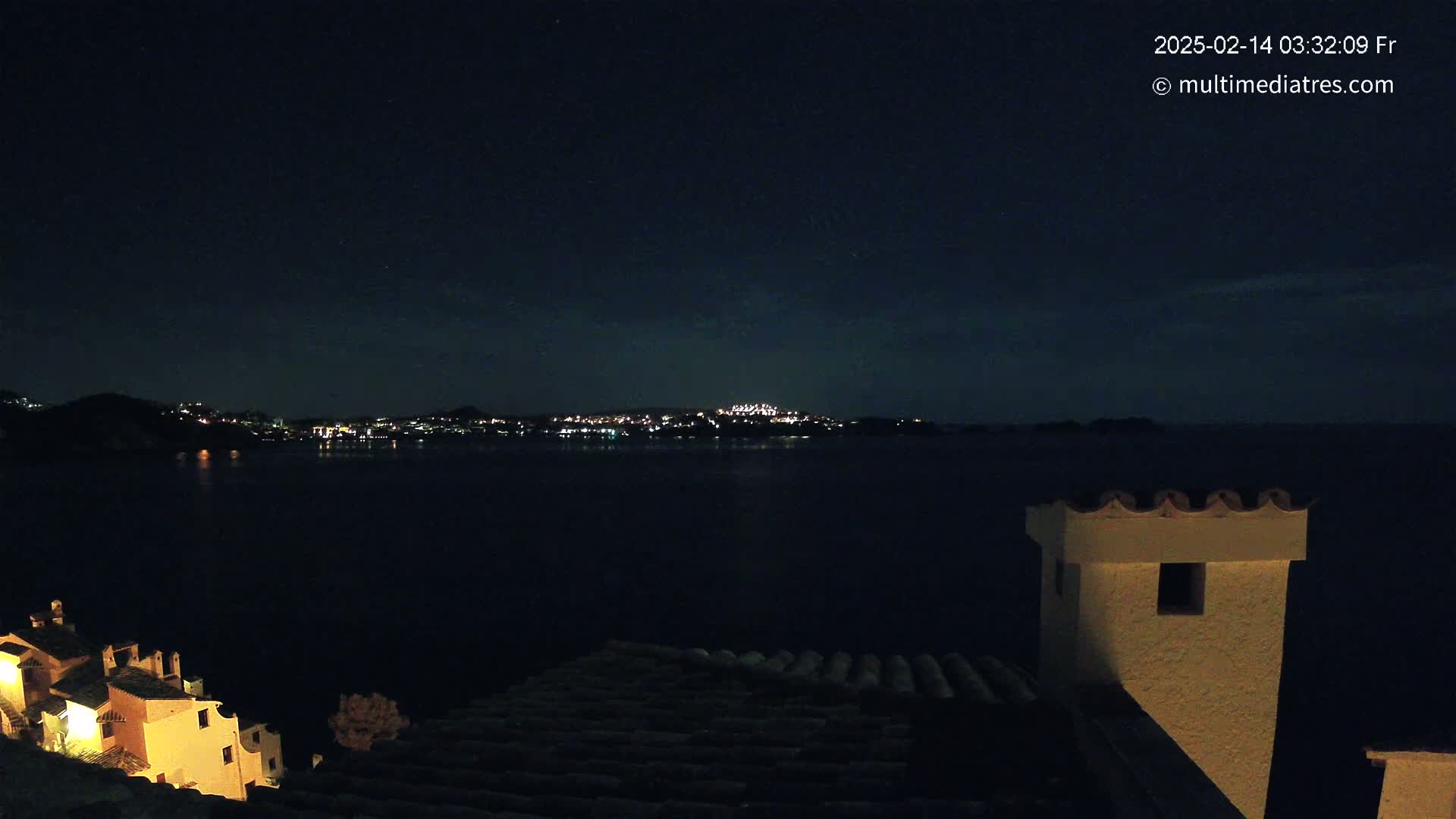 A nighttime view of a city across a dark body of water, seen from a rooftop with a chimney, under a clear, starry sky.