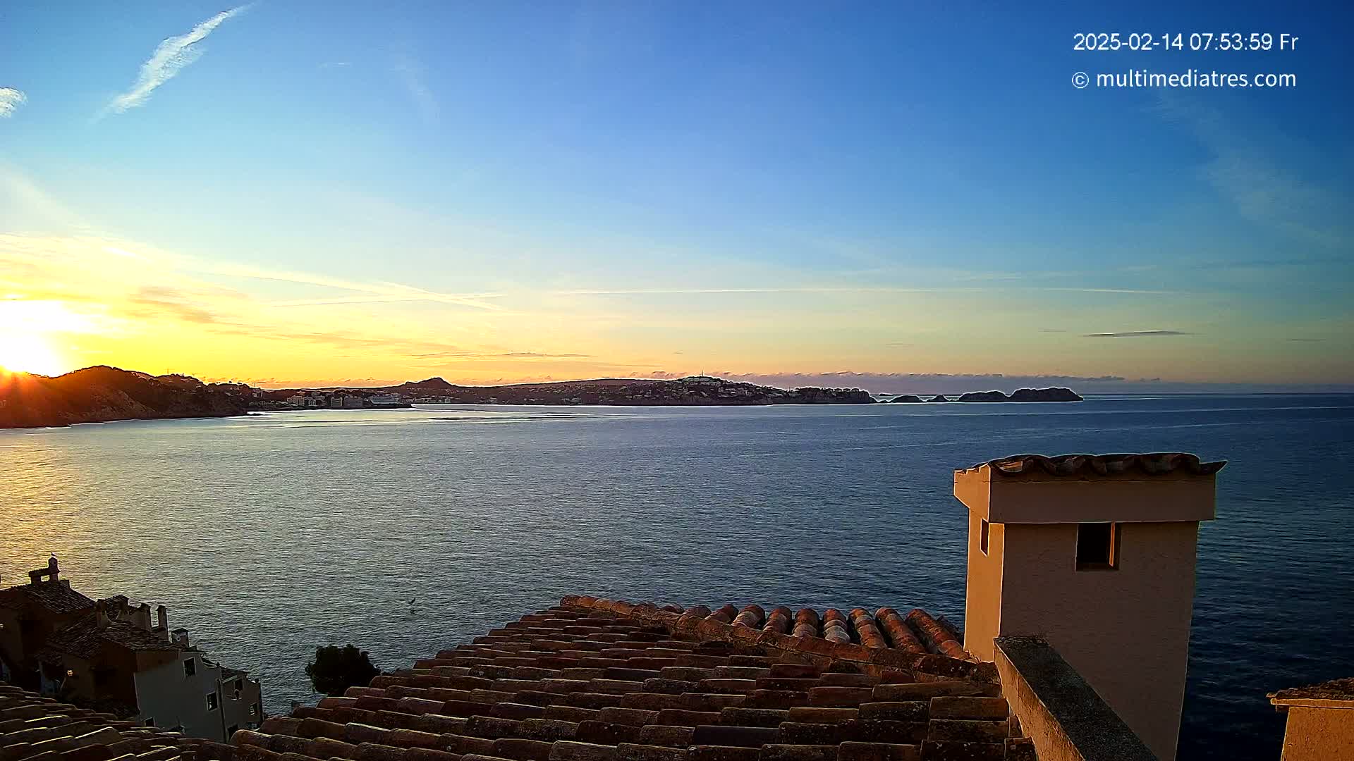 A sunrise over a calm sea, viewed from a terracotta roof with a chimney, shows a distant town nestled between hills.