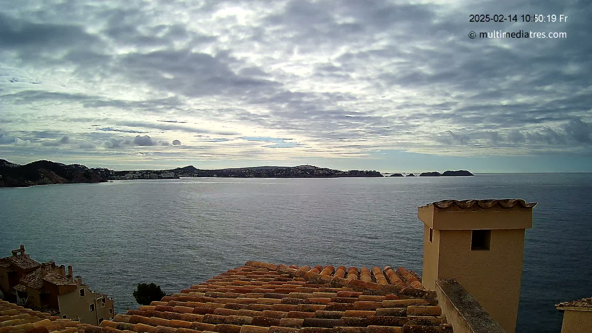 A partly cloudy sky overlooks a calm sea separating a coastal town from distant islands and hills.