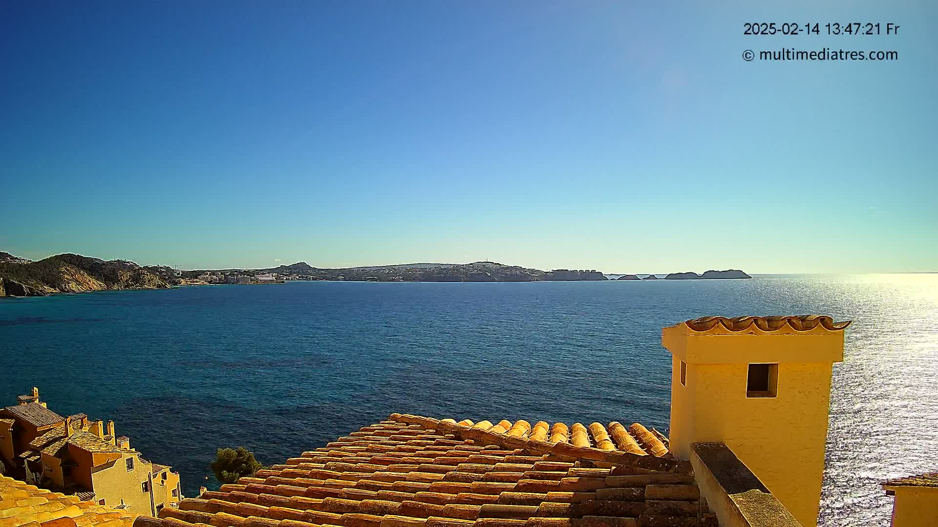 A sunlit coastal scene shows a terracotta roof overlooking a calm blue sea with distant land under a clear blue sky.