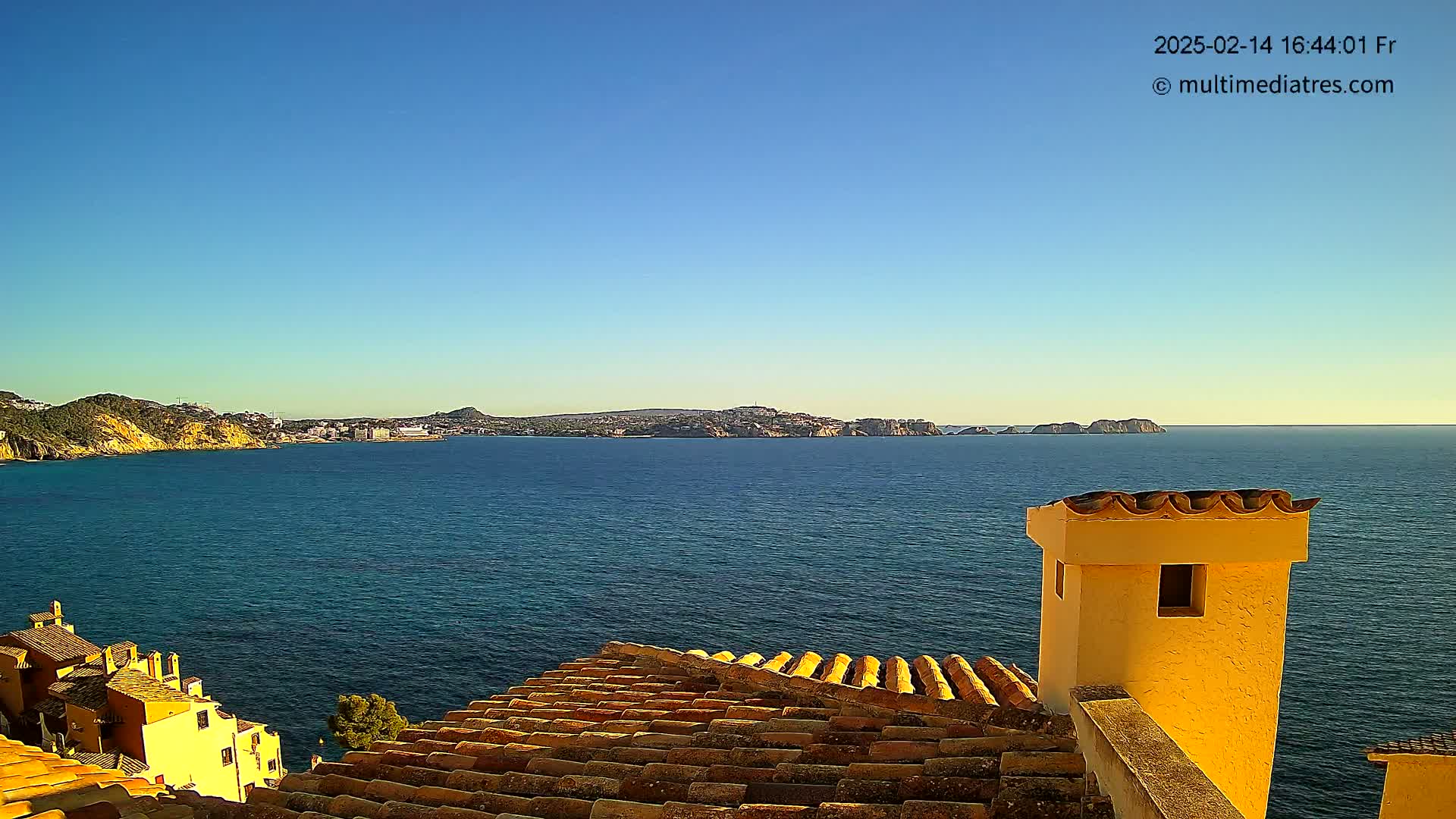 A clear, sunny day overlooks a calm blue sea from a terracotta roof, with a distant cityscape visible on the horizon and a yellow chimney in the foreground.