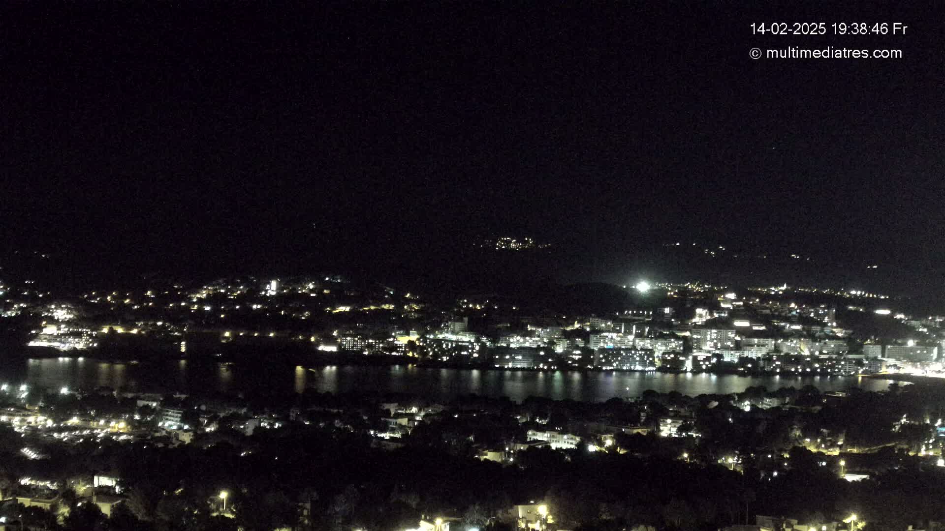 A nighttime, high-angle view shows a city illuminated by lights surrounding a body of dark water, under a clear, dark sky.