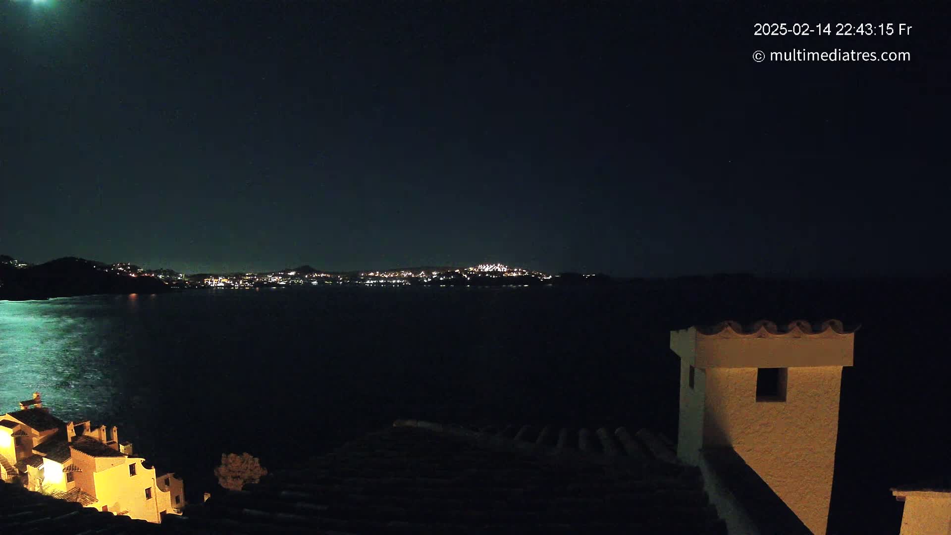 A nighttime view of a moonlit cityscape across a dark body of water, partially visible from a building's rooftop.