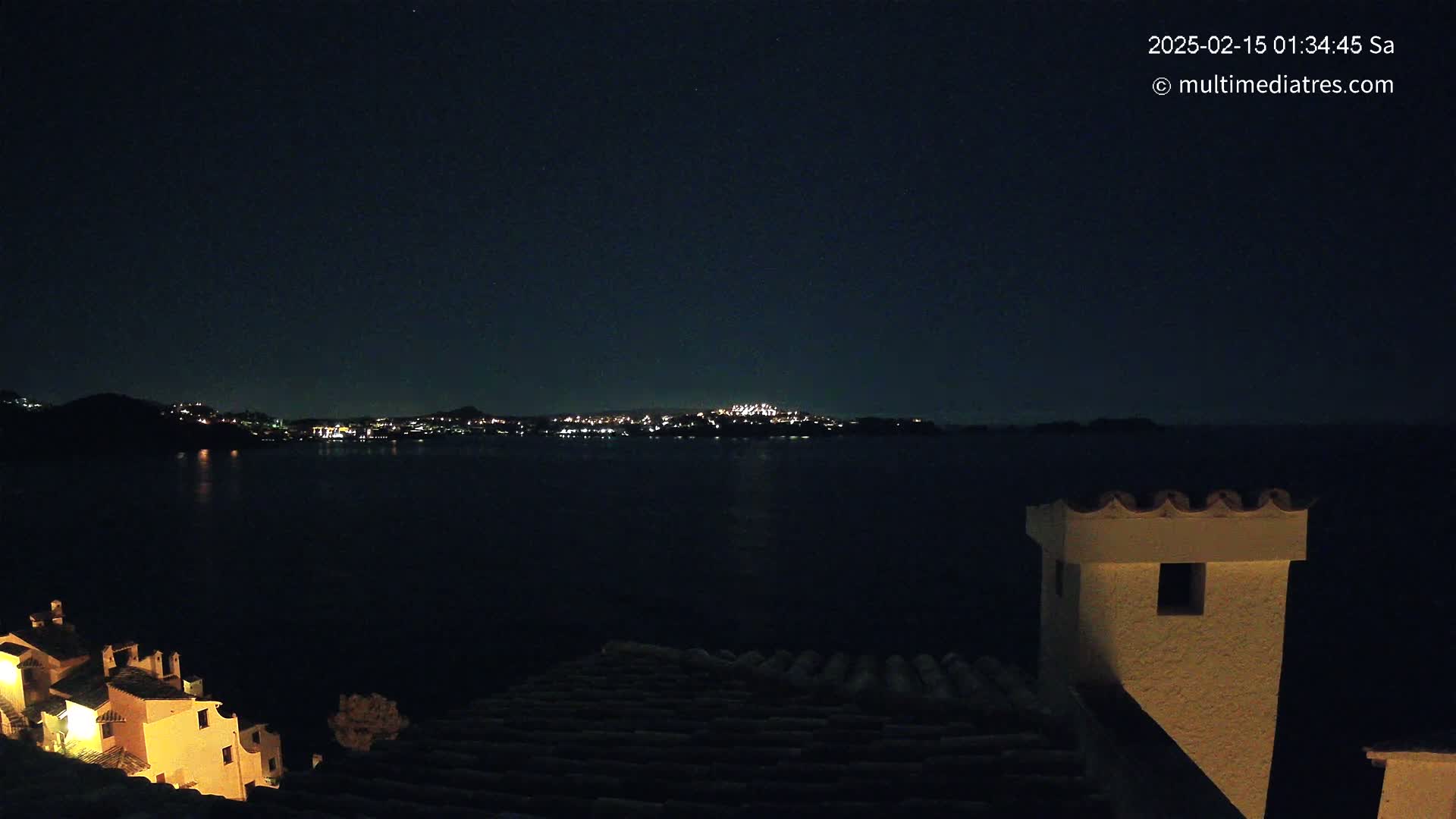 A nighttime view from a rooftop overlooking a dark body of water, with a distant city skyline illuminated by lights under a clear, starry sky.