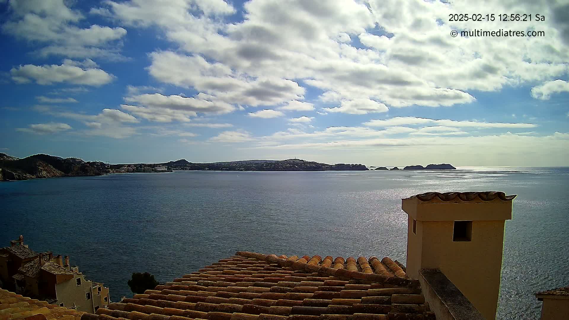 A partly cloudy sky overlooks a calm sea with distant hills and a section of a tiled roof with a chimney in the foreground.