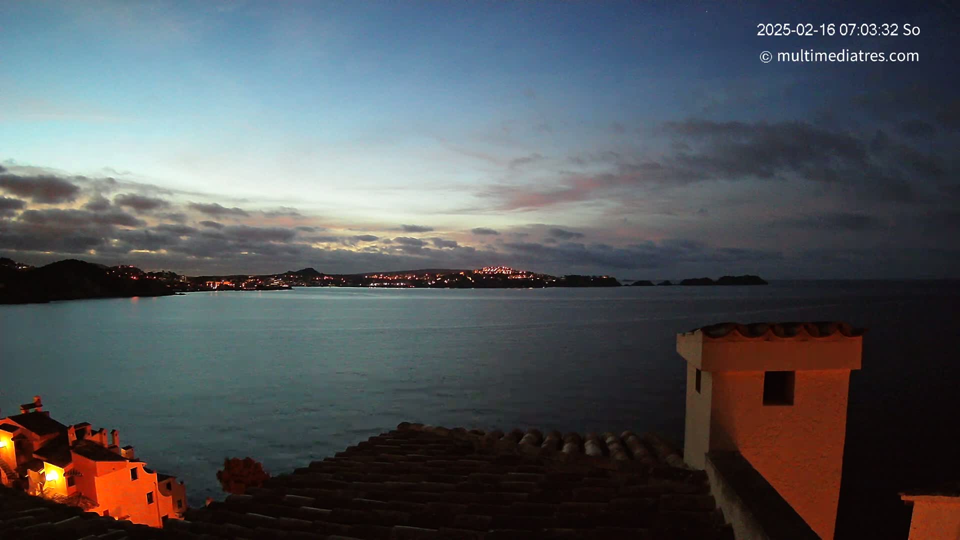 A twilight view over a calm body of water shows a town with lights across the bay, under a partly cloudy sky.