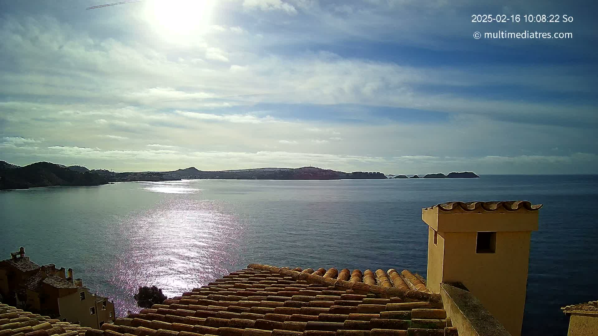 A partly sunny day overlooking a calm sea with distant snow-capped hills and a tiled rooftop in the foreground.