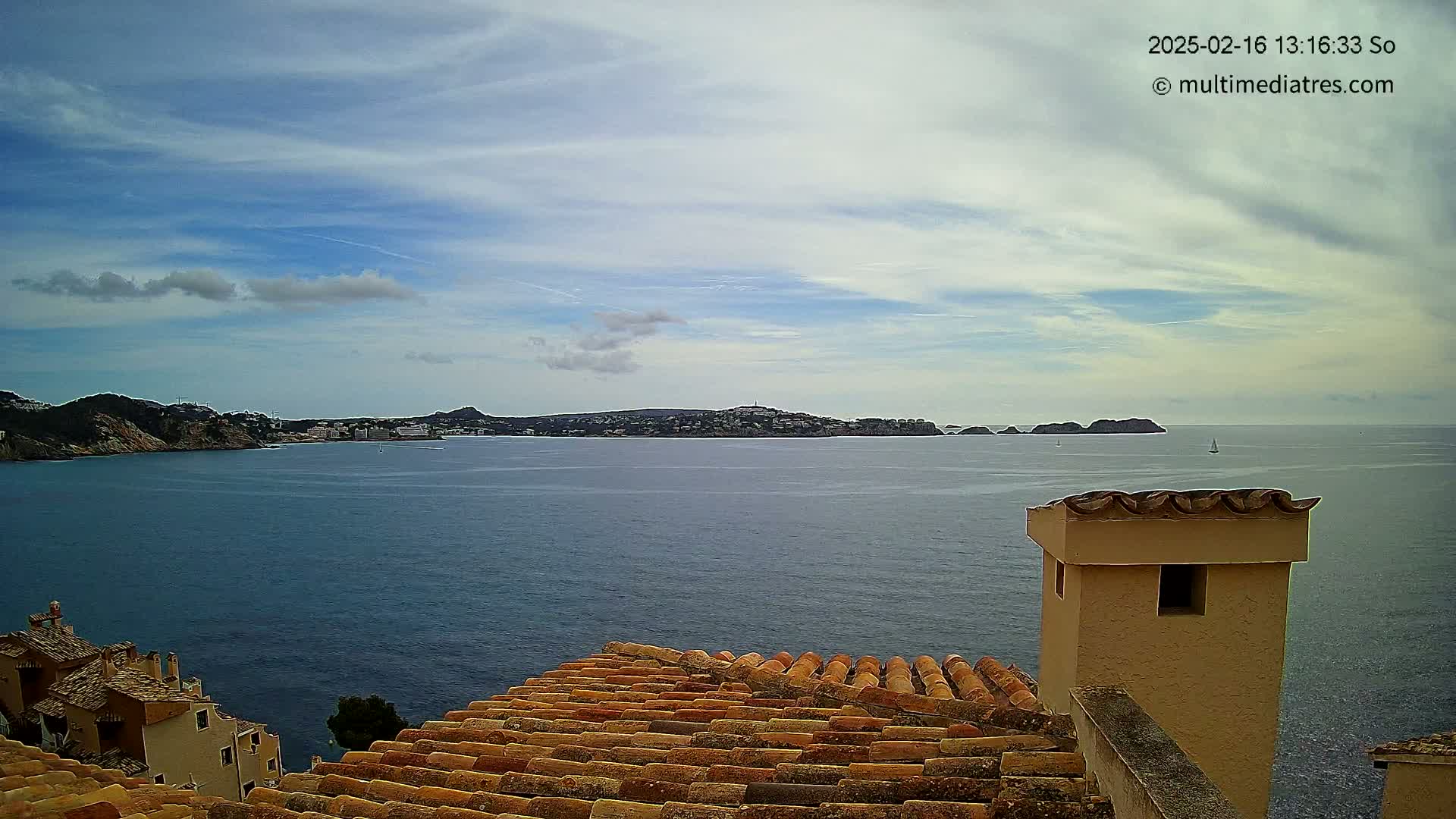 A partly cloudy sky overlooks a calm sea with a distant shoreline and buildings, viewed from a tiled roof with a chimney.