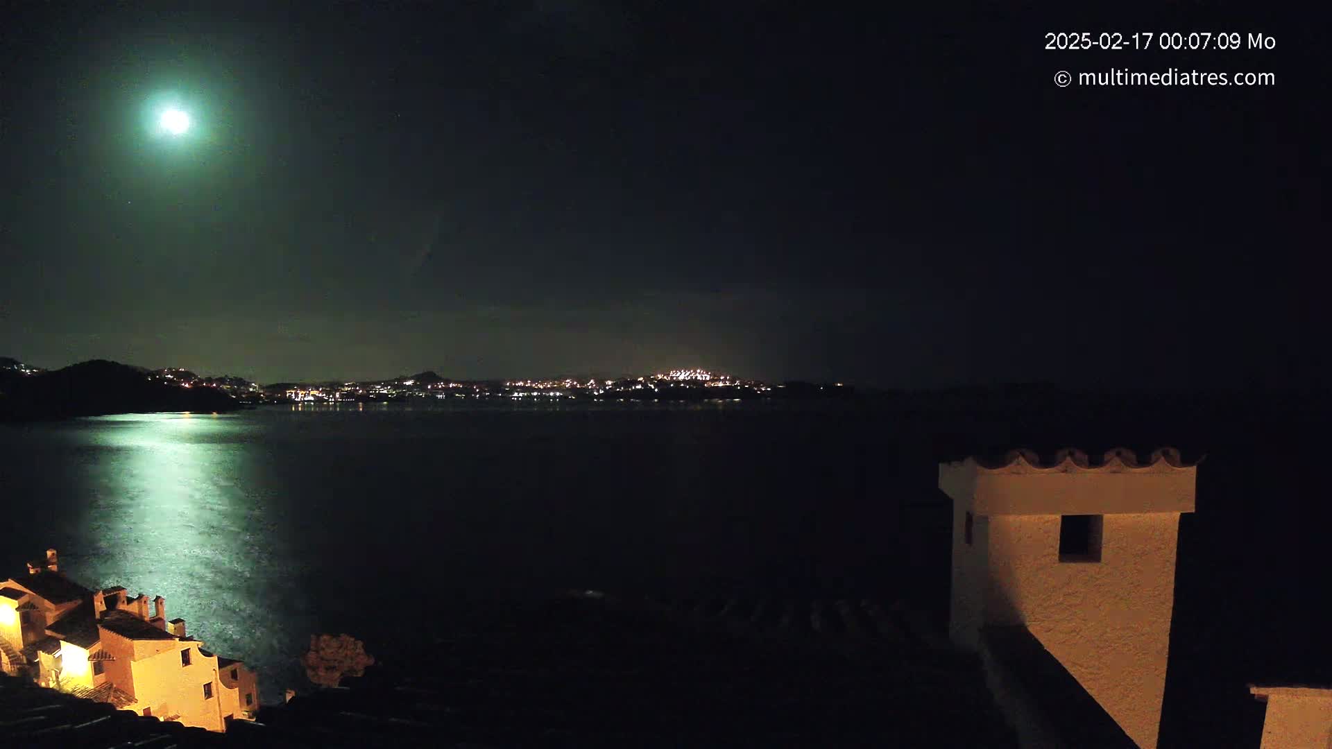 A bright moon illuminates a calm body of water reflecting its light, with a distant town glowing across the water under a clear night sky, and a portion of a building in the foreground.