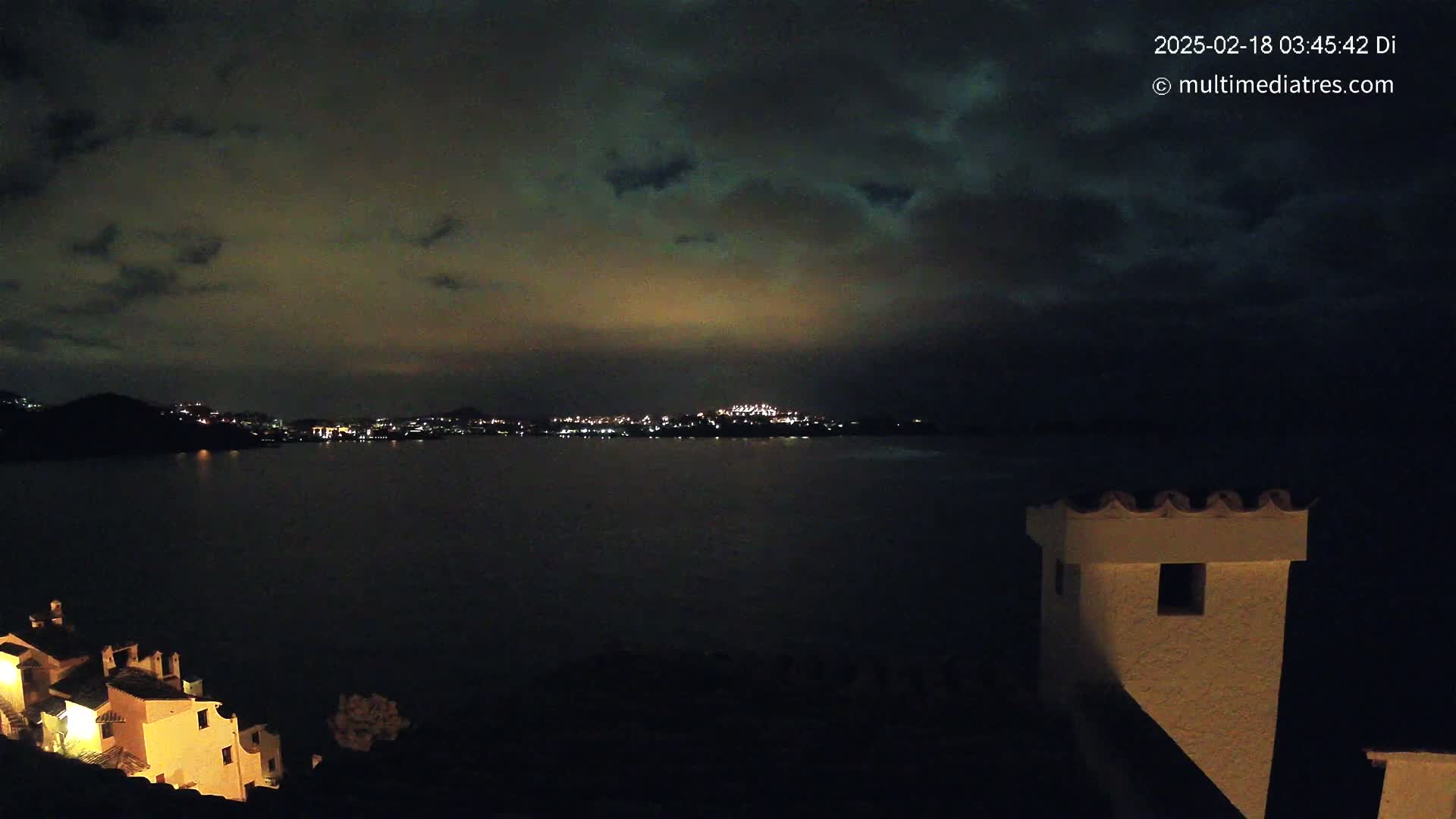 A nighttime view of a dark body of water with a distant city skyline illuminated by lights under a cloudy sky, with a portion of a building visible in the foreground.
